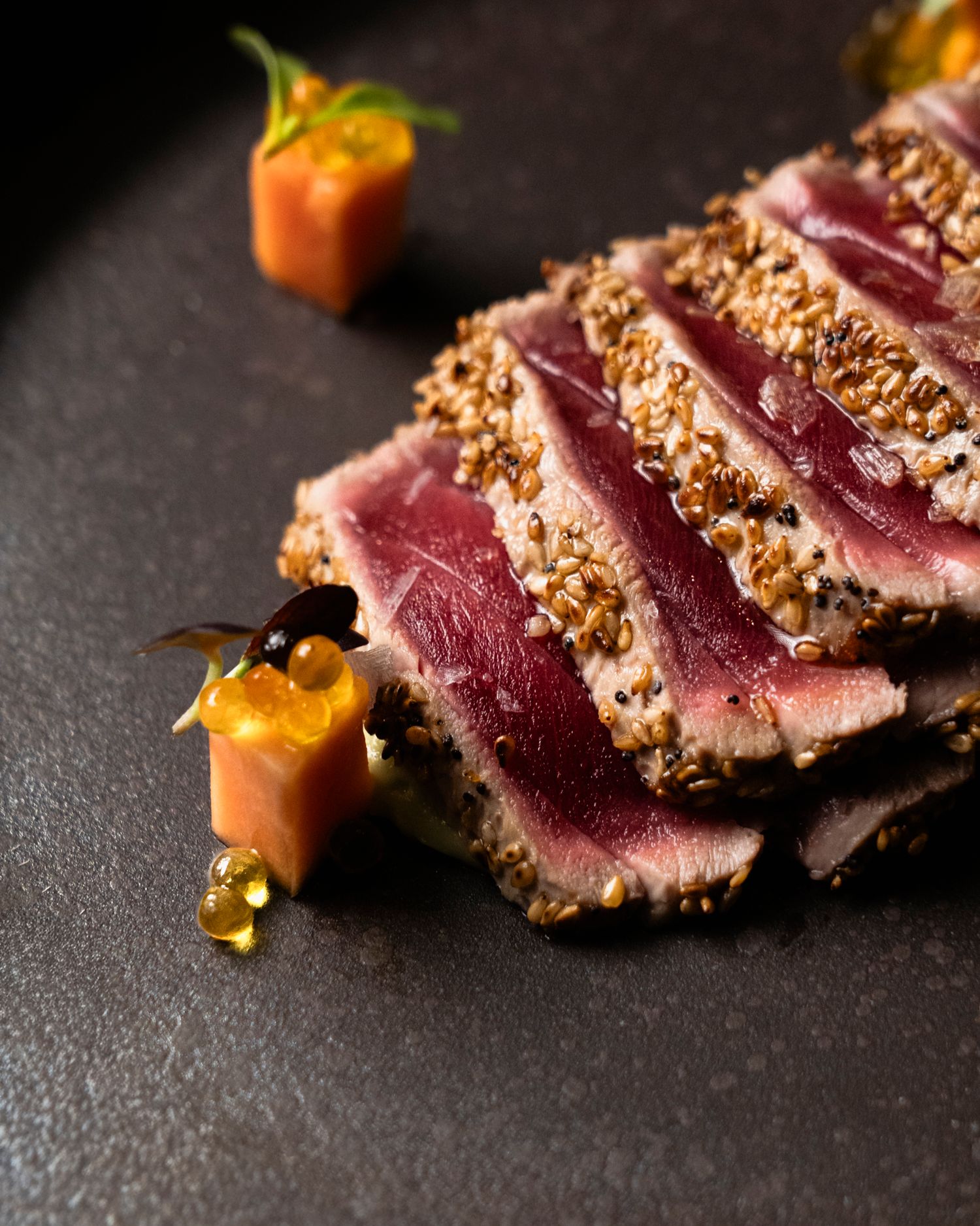 Seared sesame-crusted tuna slices with vibrant pink center, served with orange garnish on dark slate plate.