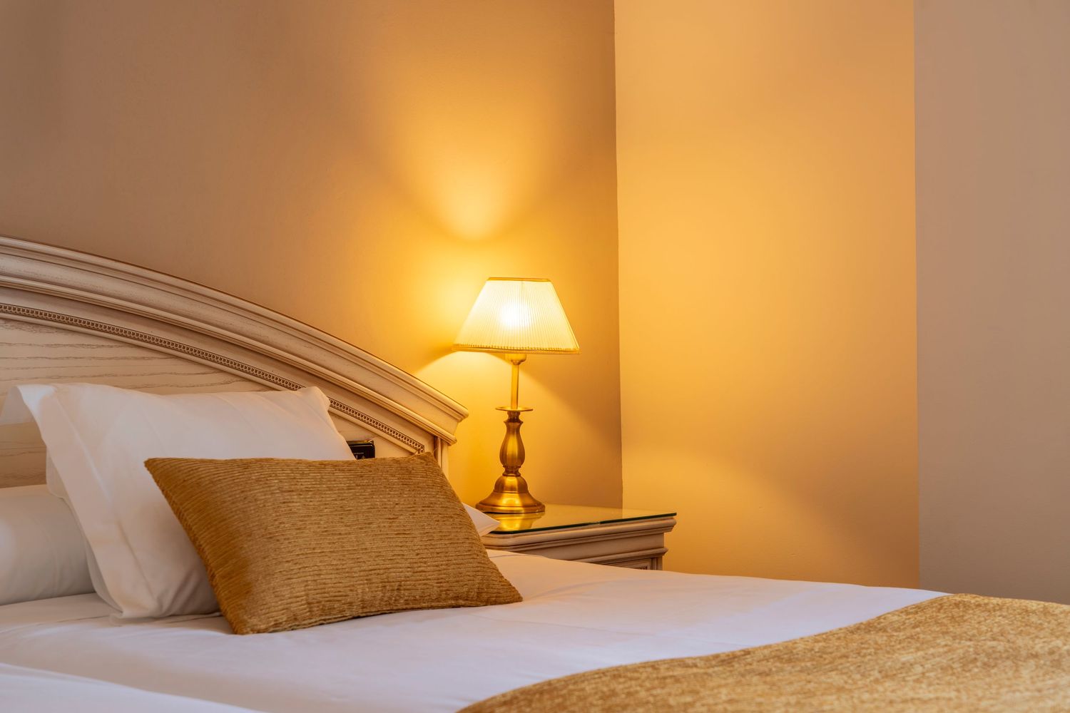 Elegant hotel bedroom with warm glowing bedside lamp, golden decorative pillow, and white bedding in a cozy setting.