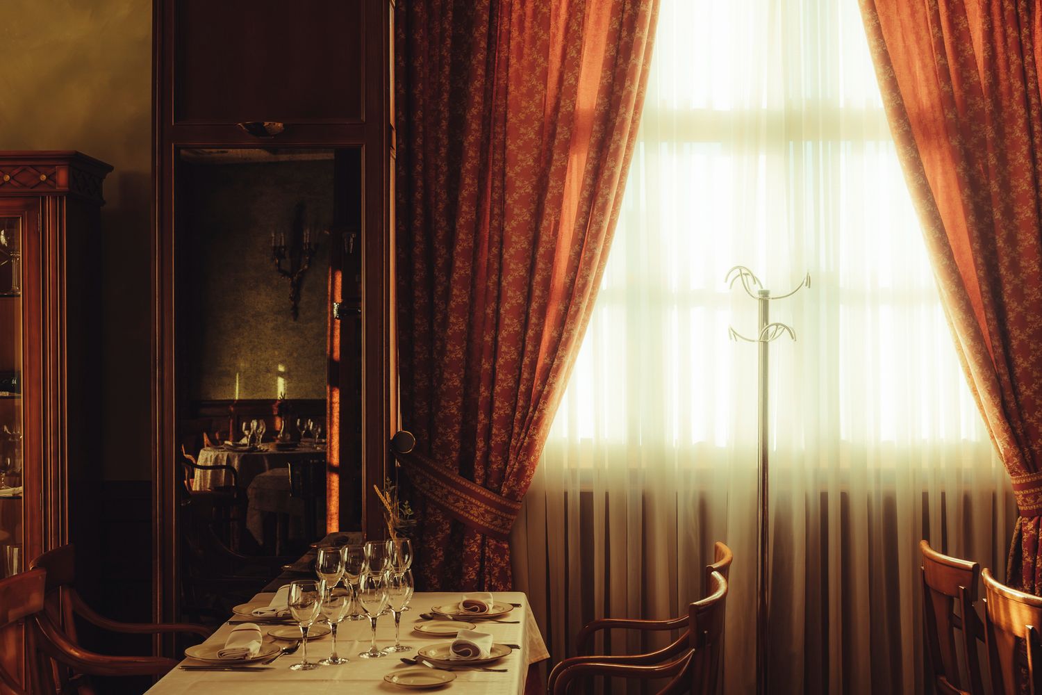 Elegant restaurant dining room with white tablecloth, wine glasses, red curtains, and warm candlelight by a large window.