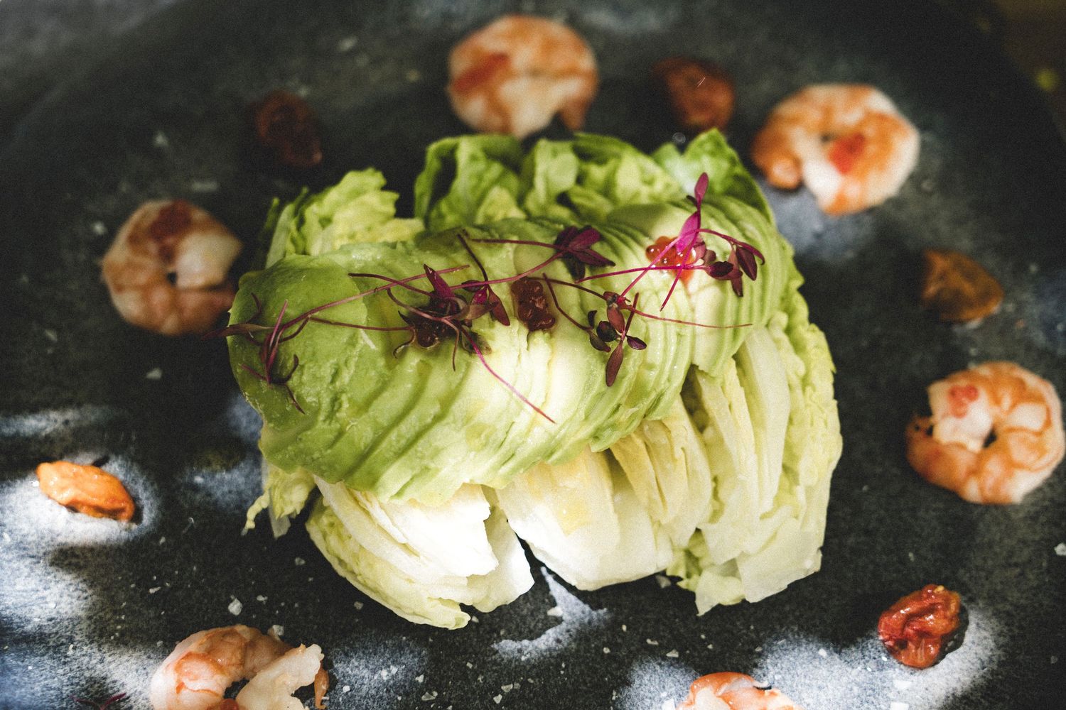 Avocado rose garnished with microgreens surrounded by sautéed shrimp on a dark ceramic plate.