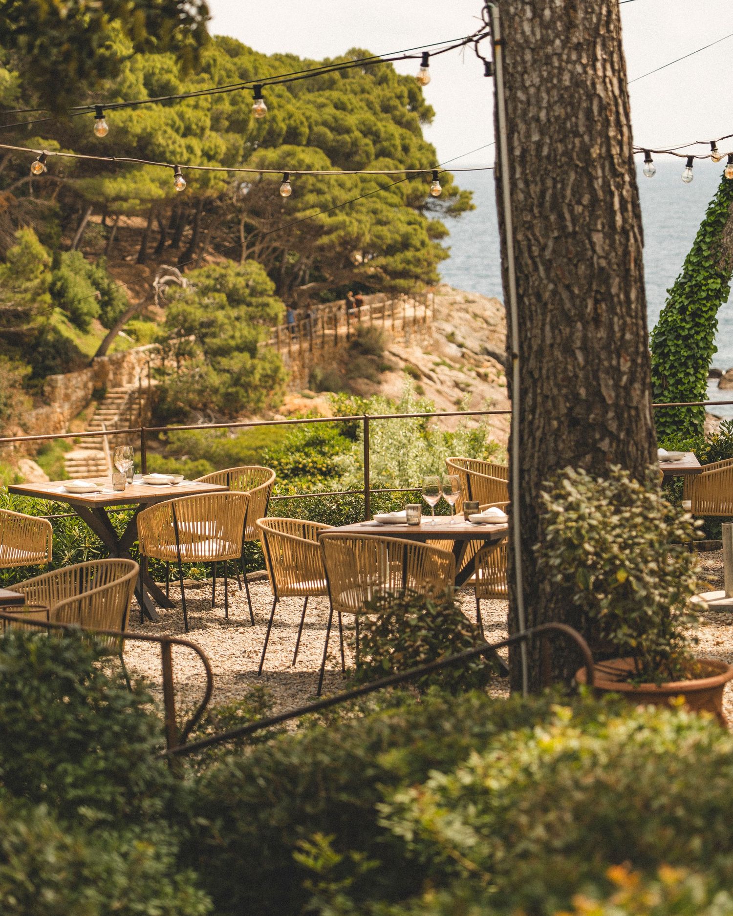 Charming outdoor restaurant terrace with rattan chairs, string lights, and scenic coastal views among lush greenery.