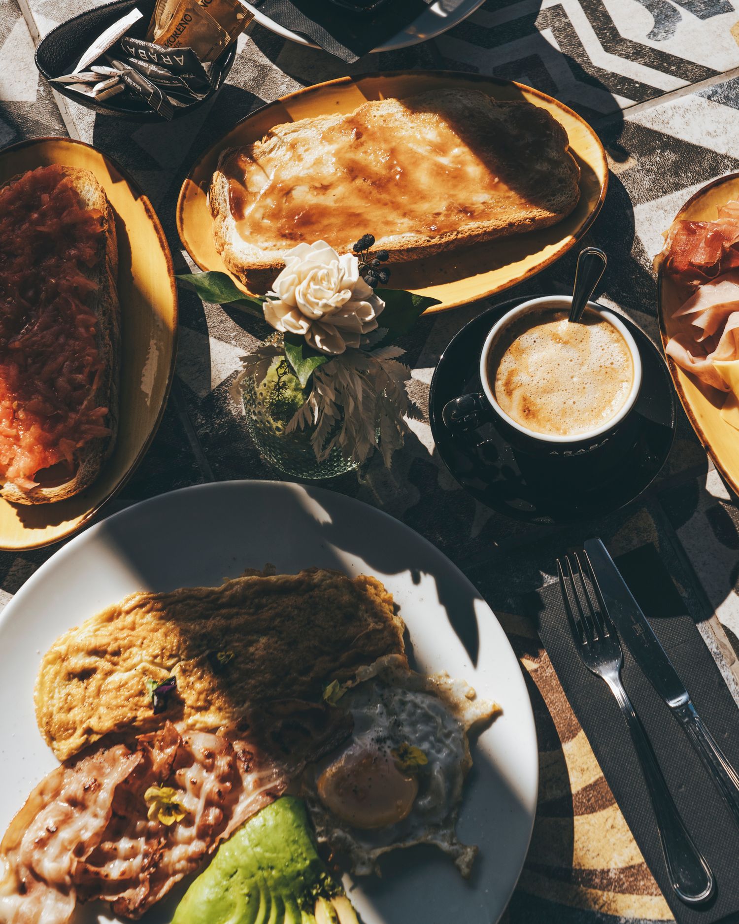 Overhead view of a breakfast spread with eggs, bacon, toast, and coffee on a sunlit restaurant table.