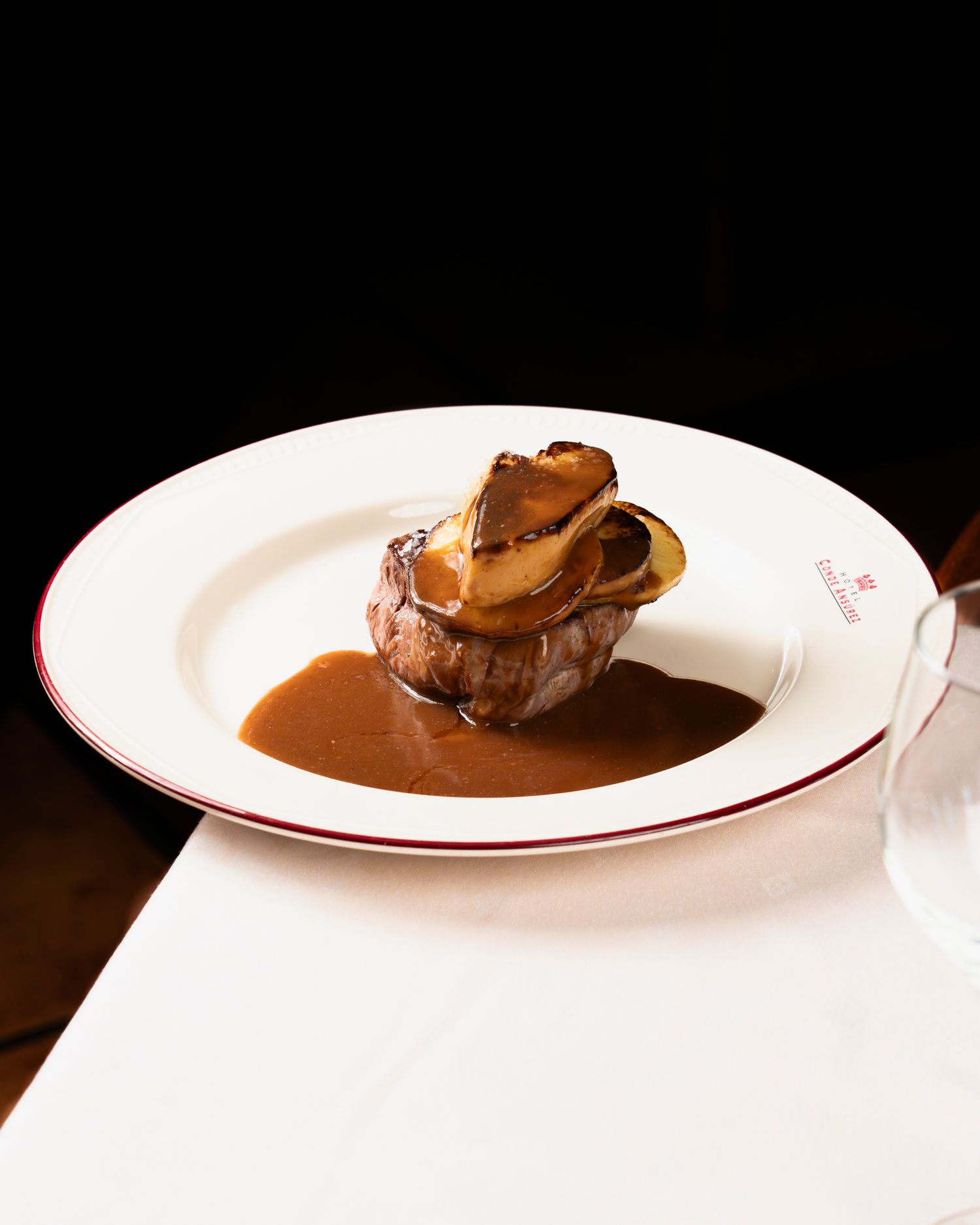 Elegant filet mignon topped with seared foie gras in rich brown sauce served on a white plate with red trim.