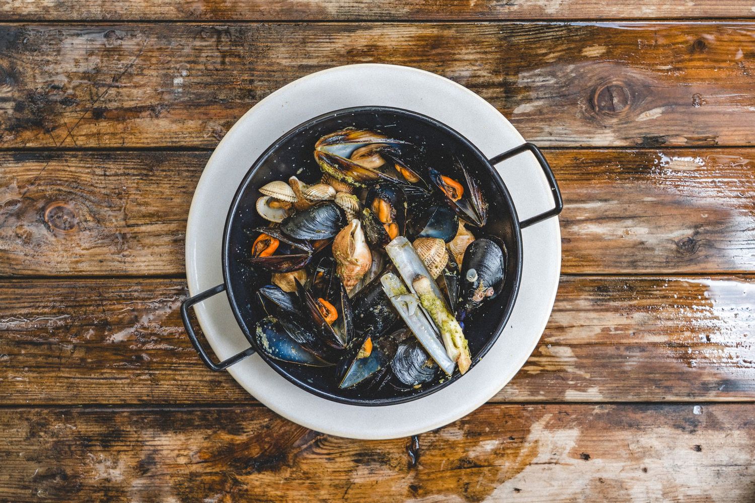 Steamed mussels in a black pan served on a white plate on a rustic wooden table.