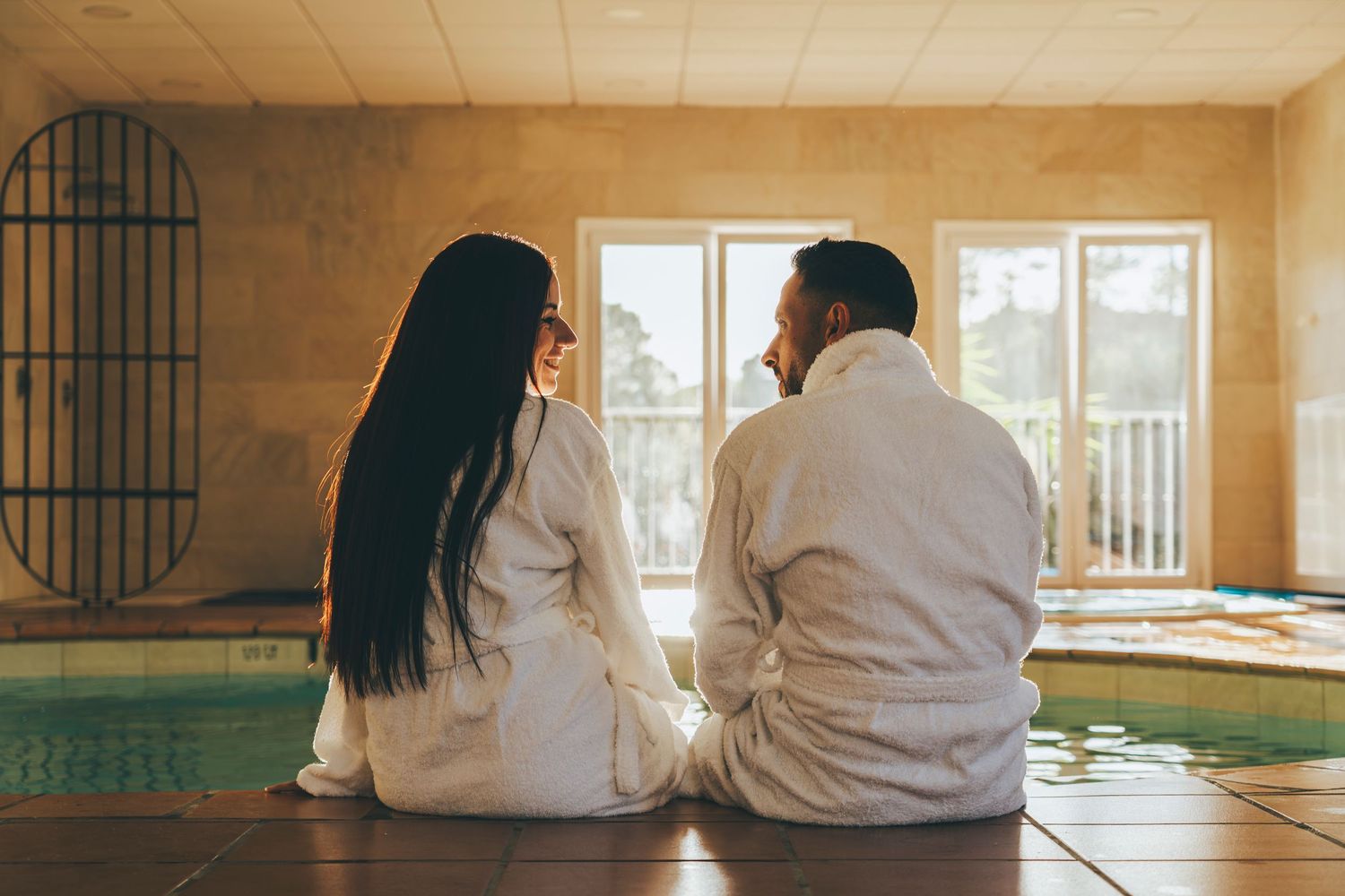 Couple in white robes sitting together by an indoor pool in a luxury spa retreat.