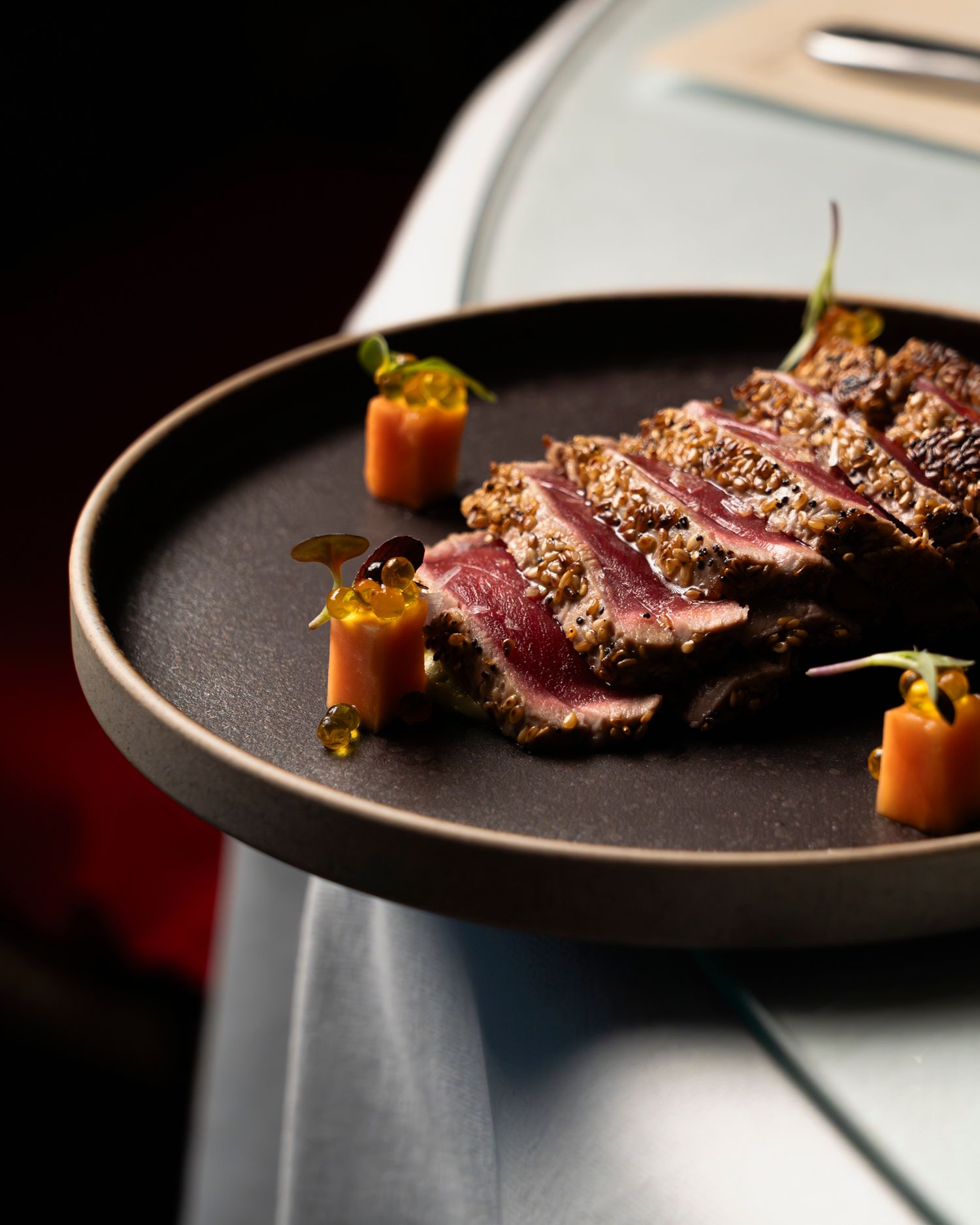 Sliced seared beef with herb crust, microgreens, and glazed carrot garnish on a dark ceramic plate.
