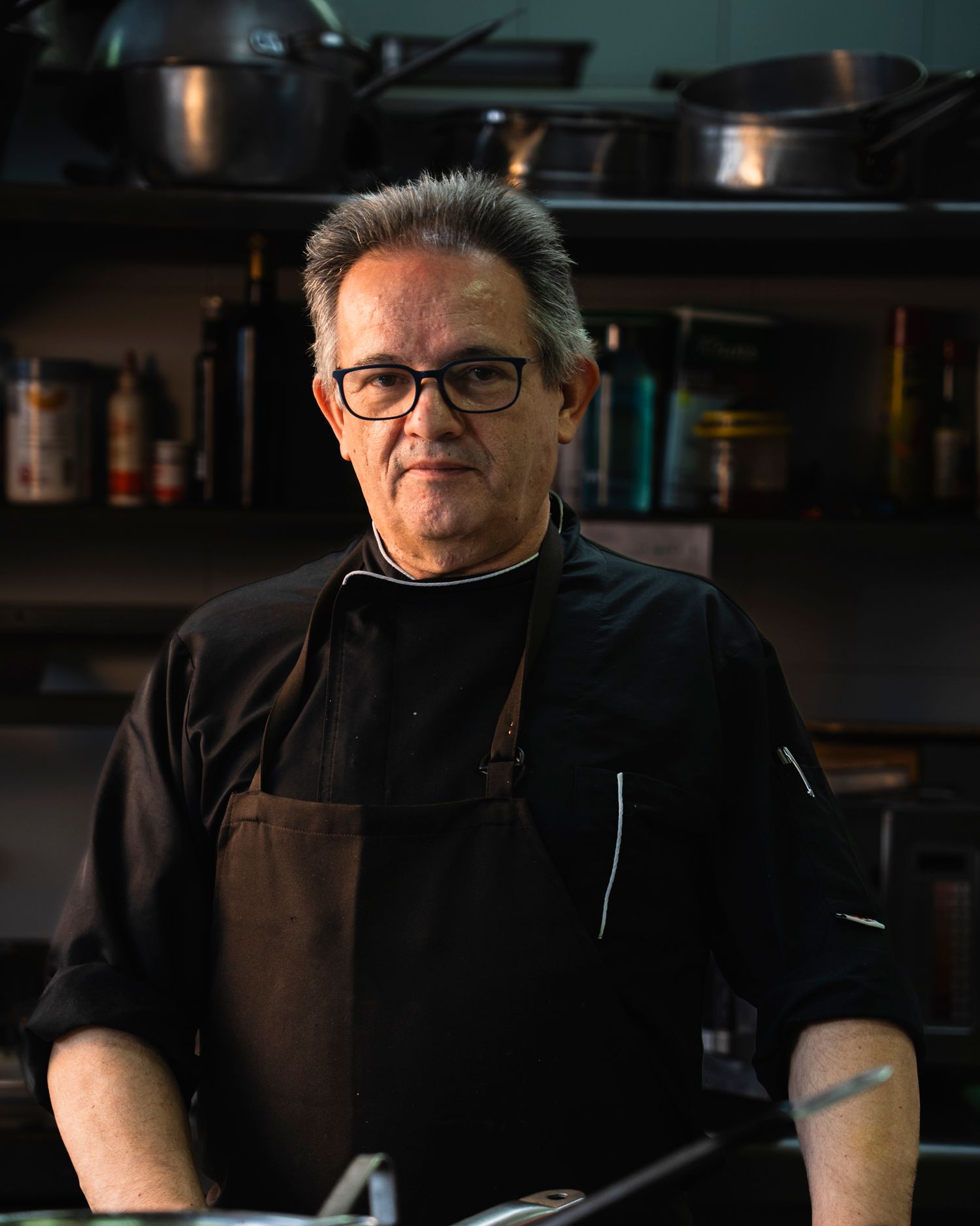 A seasoned chef in black jacket and brown apron stands confidently in a professional kitchen.