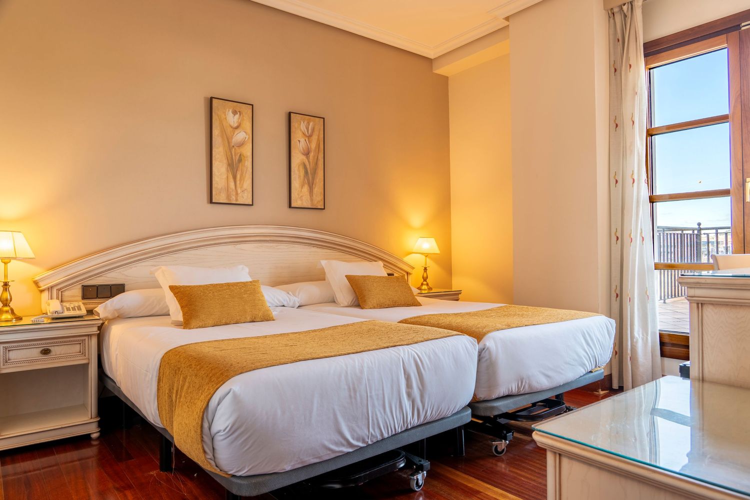 Bright hotel room with twin beds, gold accent bedding, white headboards, hardwood floors, and window with curtains.
