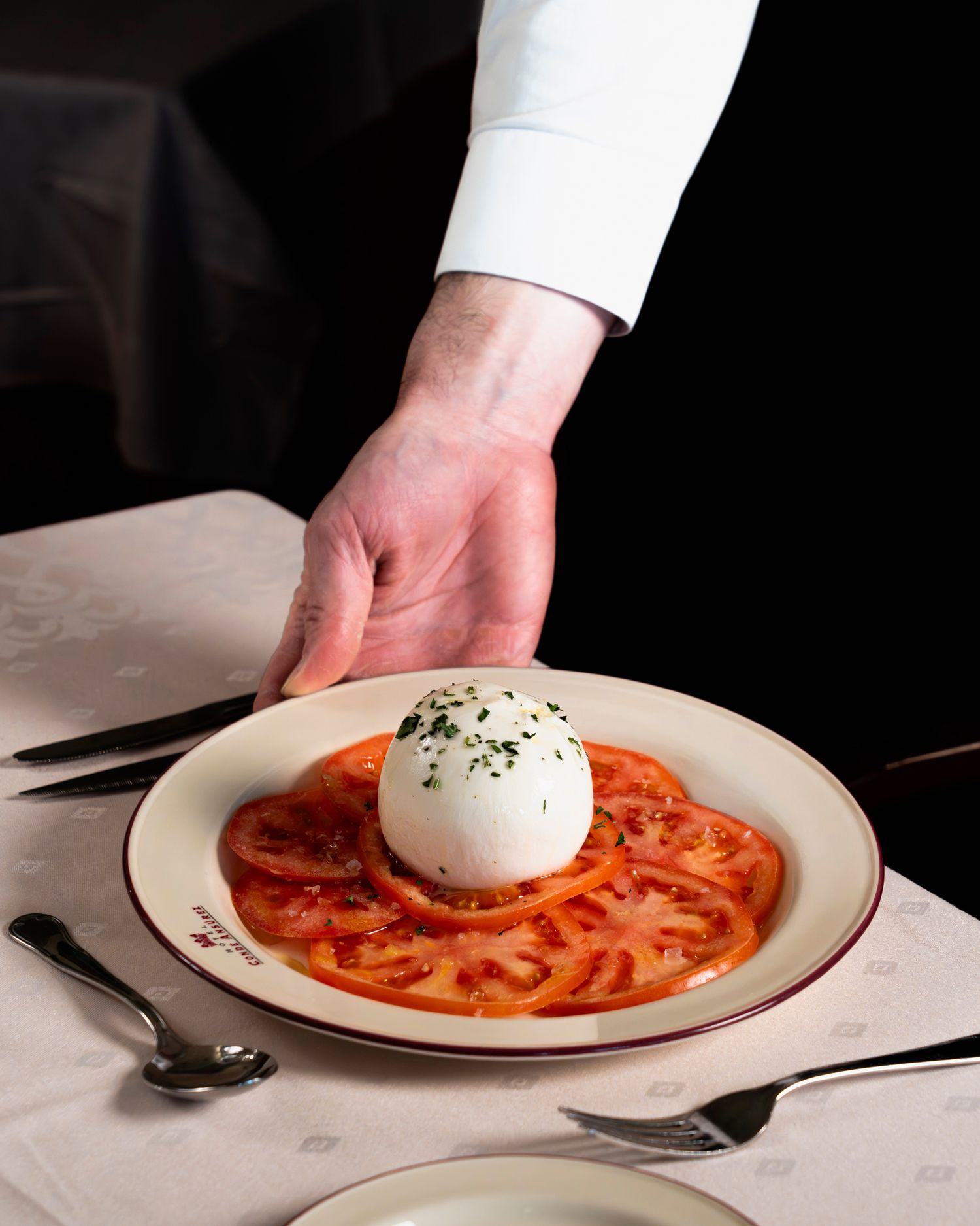 Chef presenting burrata cheese on sliced heirloom tomatoes on a white plate with red trim in a fine dining setting.