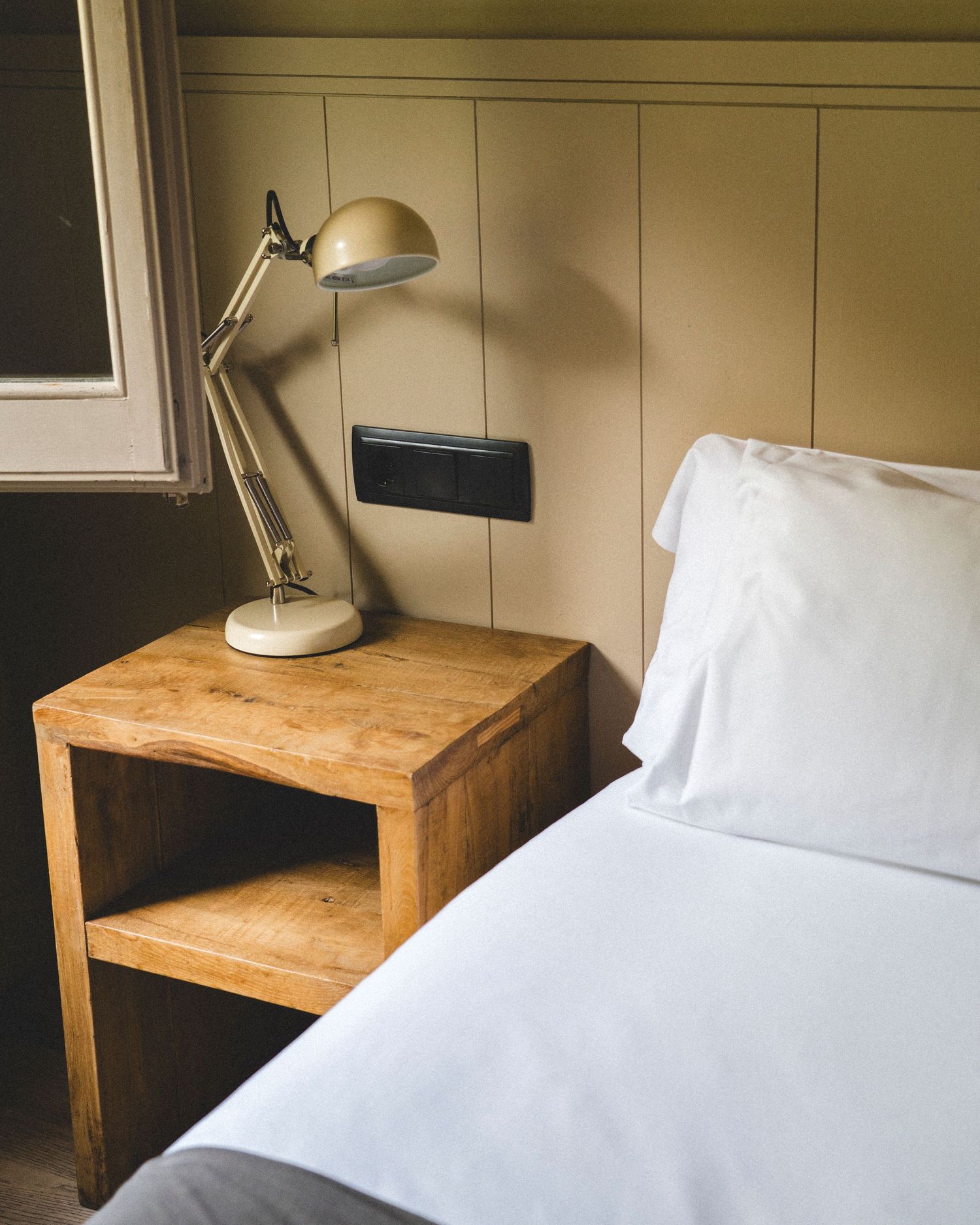 Minimalist hotel bedroom detail featuring wooden nightstand with vintage adjustable desk lamp beside white pillow bed.