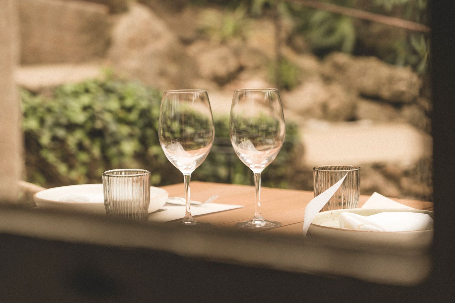 Elegant restaurant table setting with two wine glasses, folded napkins, and candle viewed through a window.
