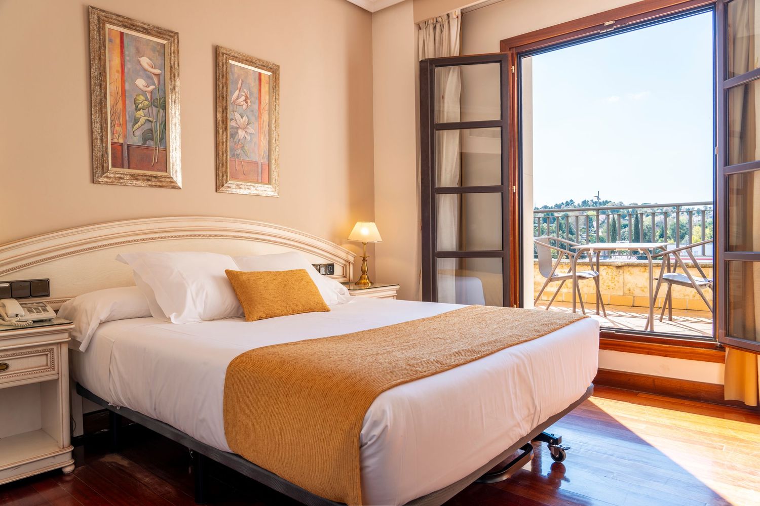 Classic hotel room with white bedding, orange accents, wooden furniture, and open balcony with scenic view.