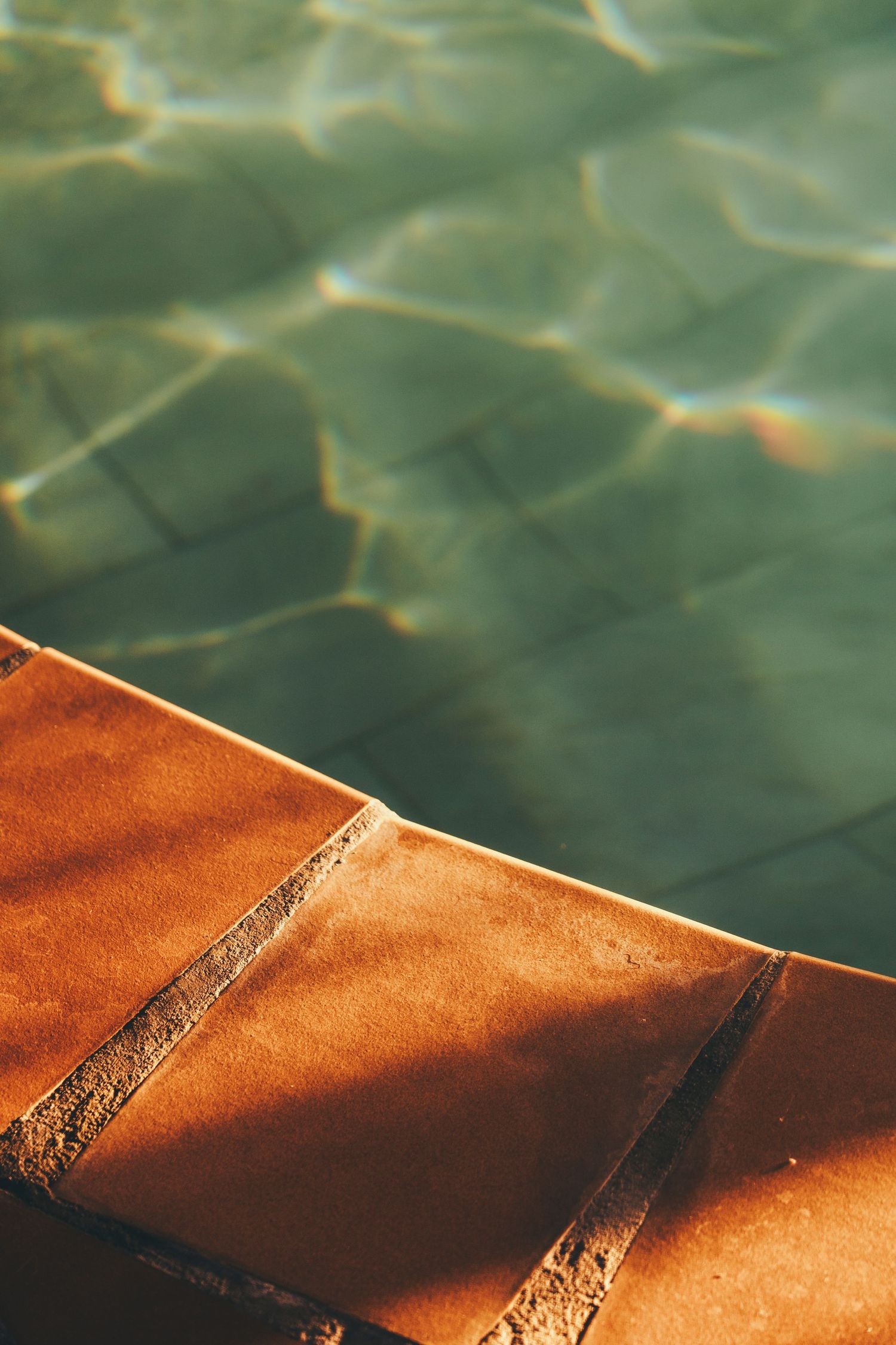 Sunlit terracotta pool steps beside shimmering turquoise water with light reflections.