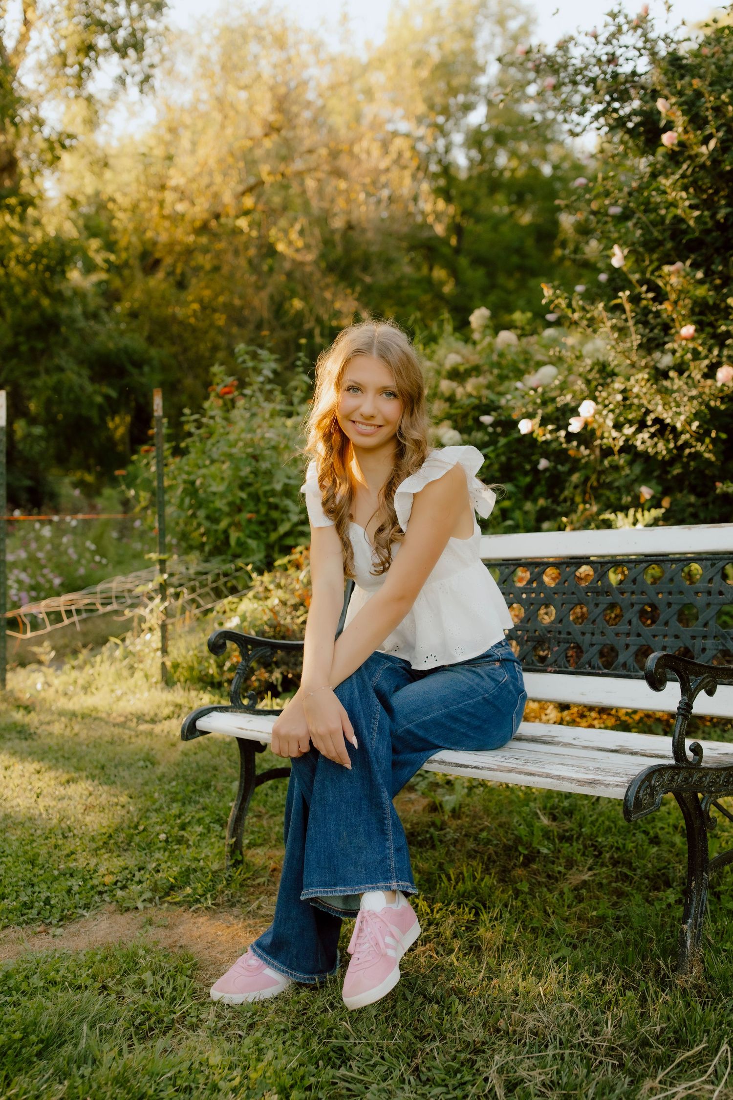 Smiling blonde girl in white top and jeans sitting on ornate bench in sunny garden setting.