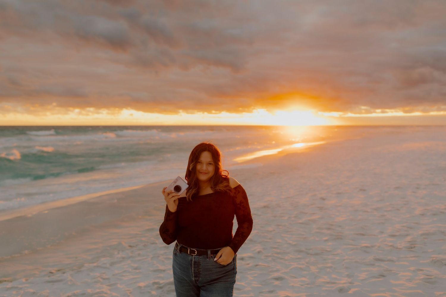Woman in dark sweater holding camera on sandy beach during vibrant golden sunset over ocean.