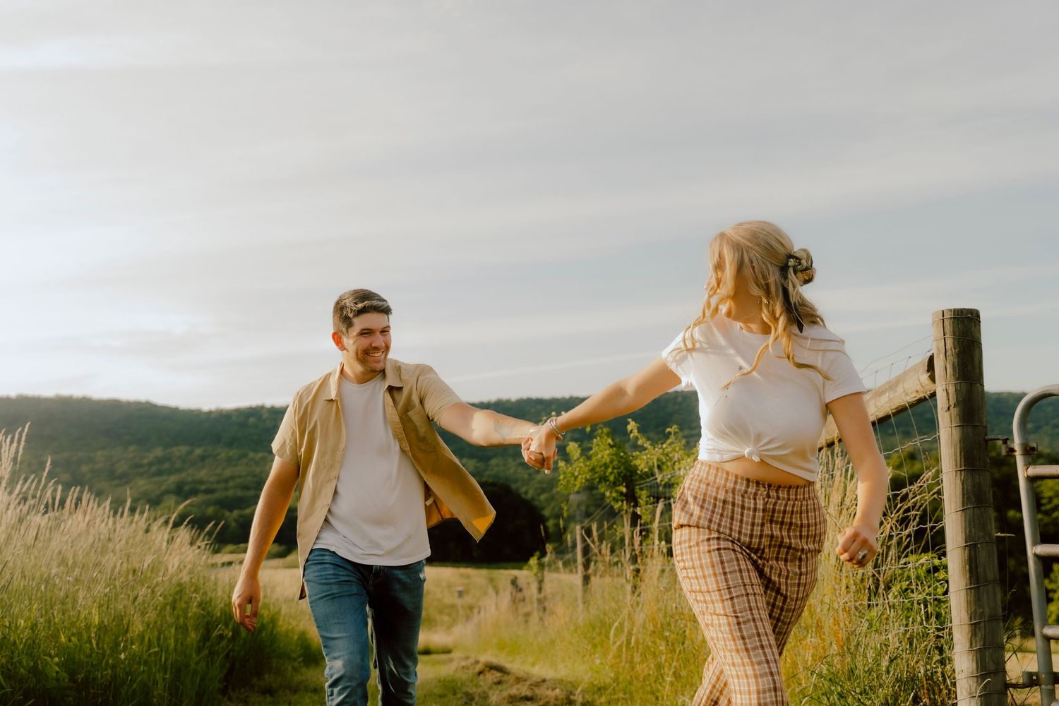 Couple holding hands walking through a scenic vineyard field with rolling green hills in the background on a sunny day.