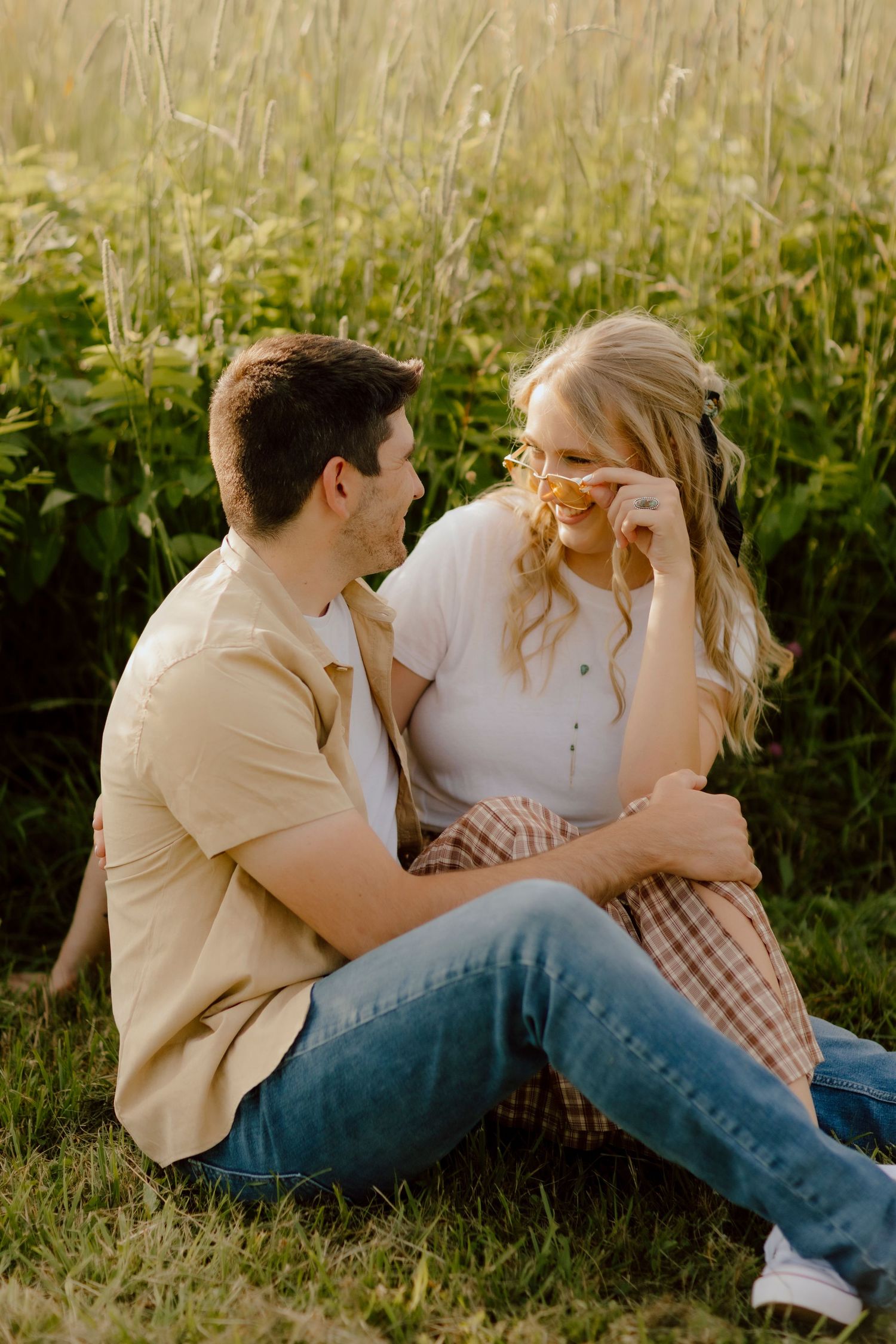 A couple sits together in a sunny field, smiling intimately as she playfully adjusts her yellow sunglasses.