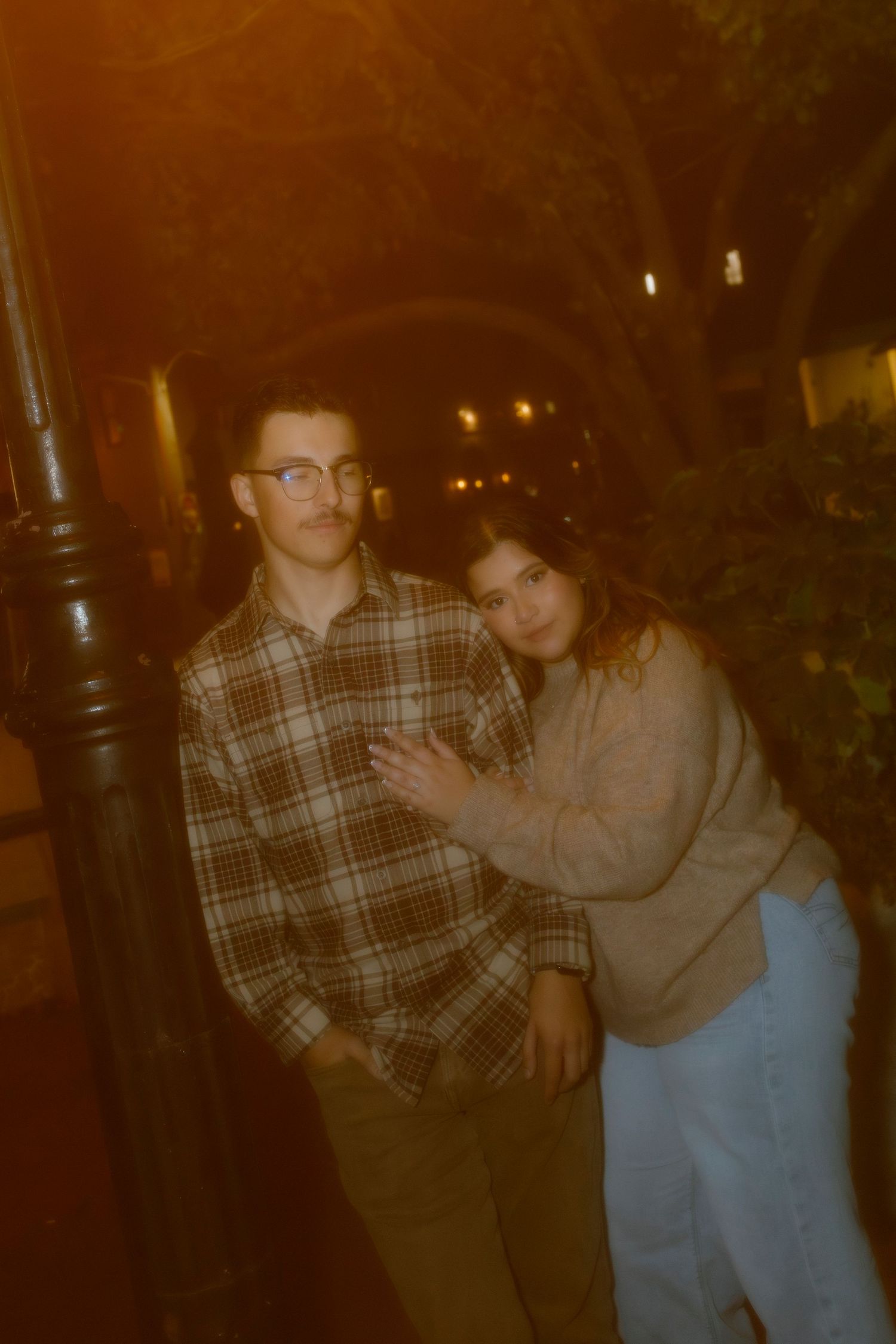 Couple leaning against a lamppost together on a warmly lit city street at night.