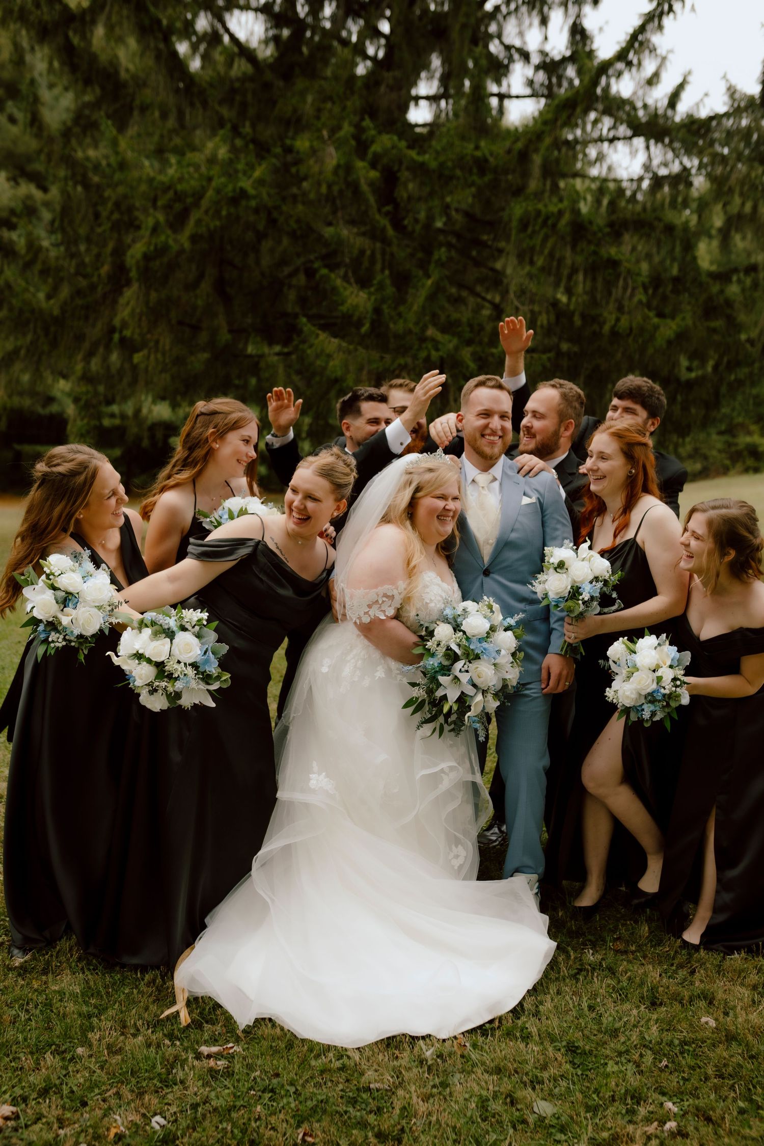 Wedding party celebrates with bride in white gown and groom in blue suit, bridesmaids in black dresses holding white bouquets.