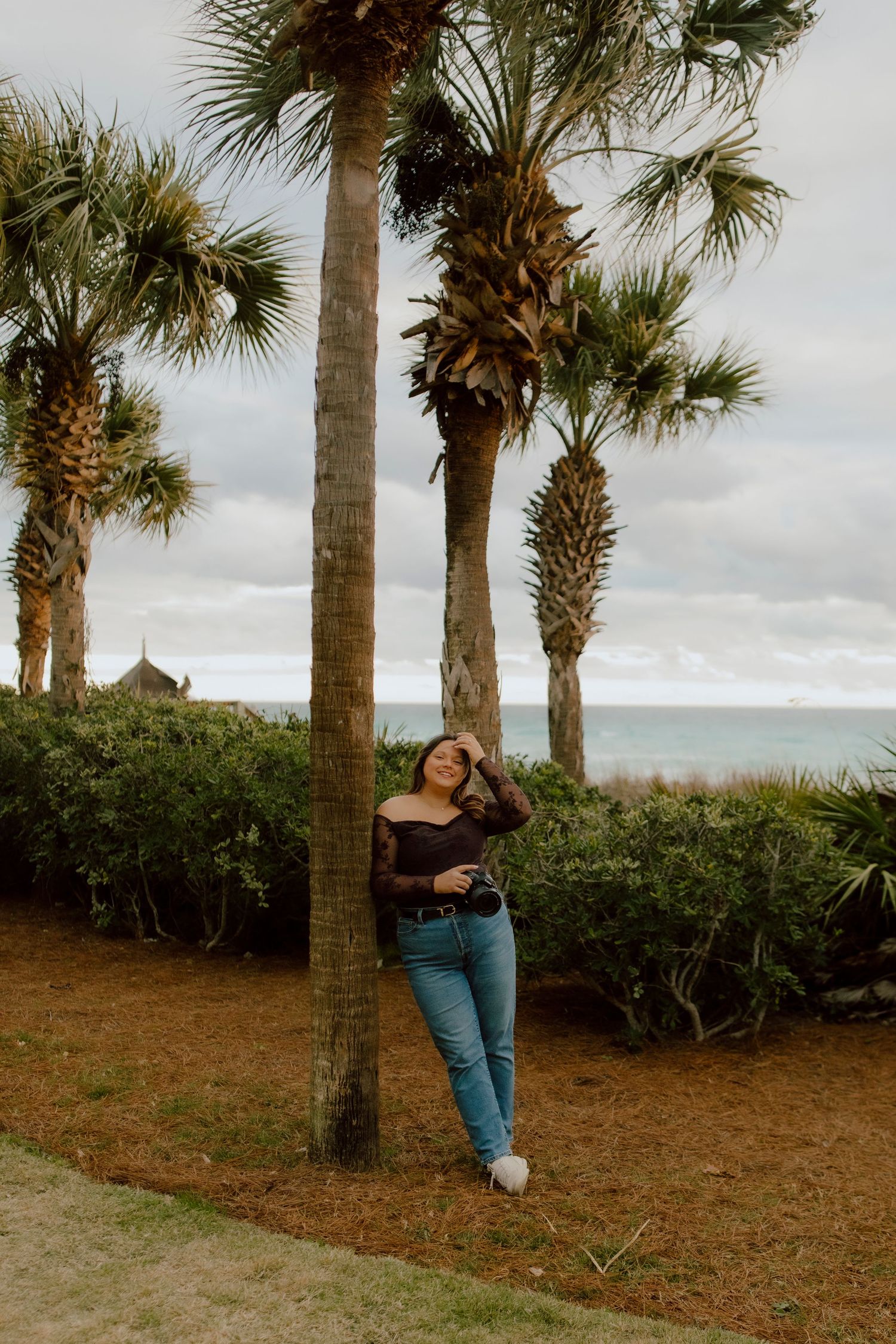 Woman in dark lace top and jeans leaning against palm tree near ocean beach on cloudy day.