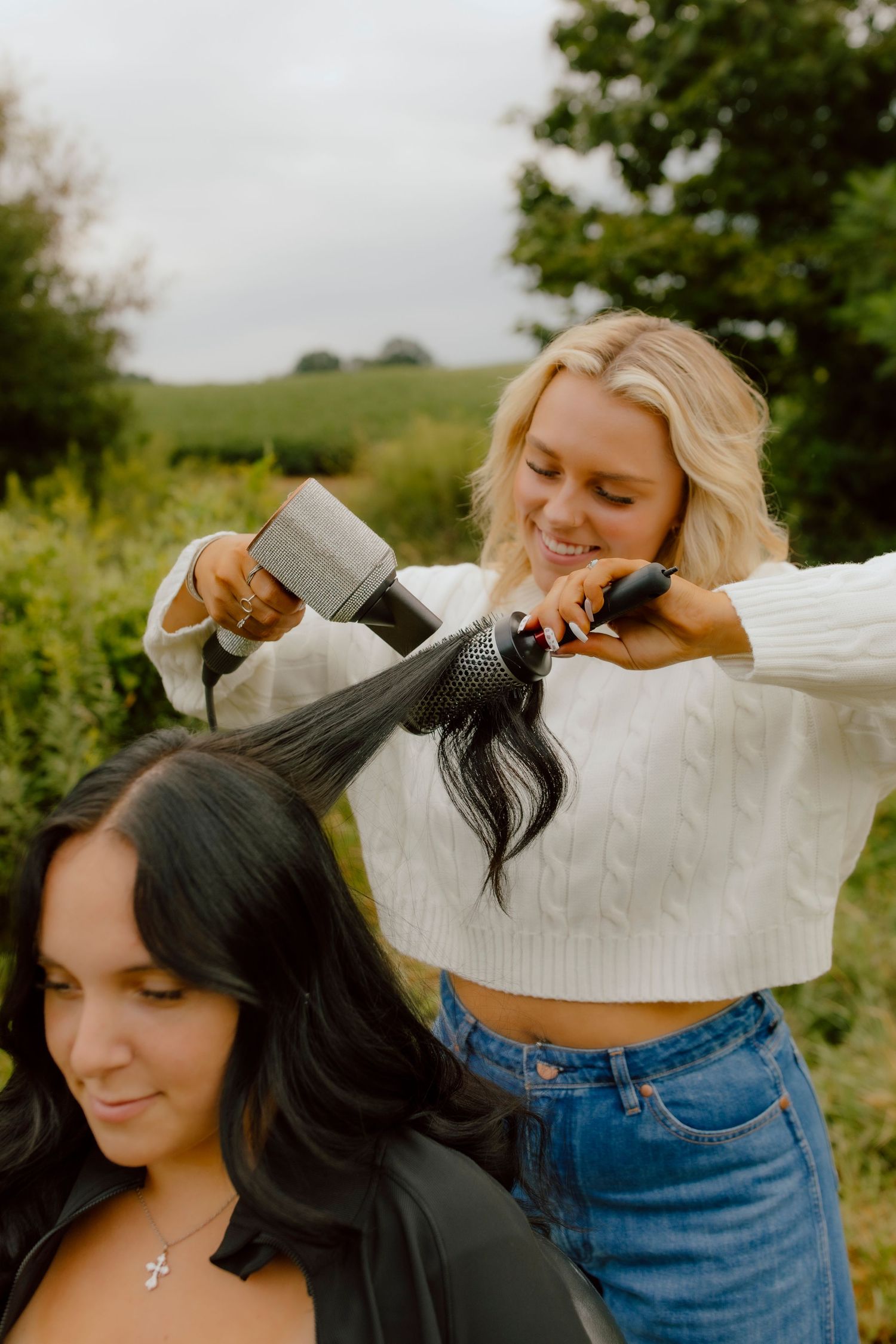 Blonde hairstylist blow-drying long dark hair outdoors with green hills in the background.