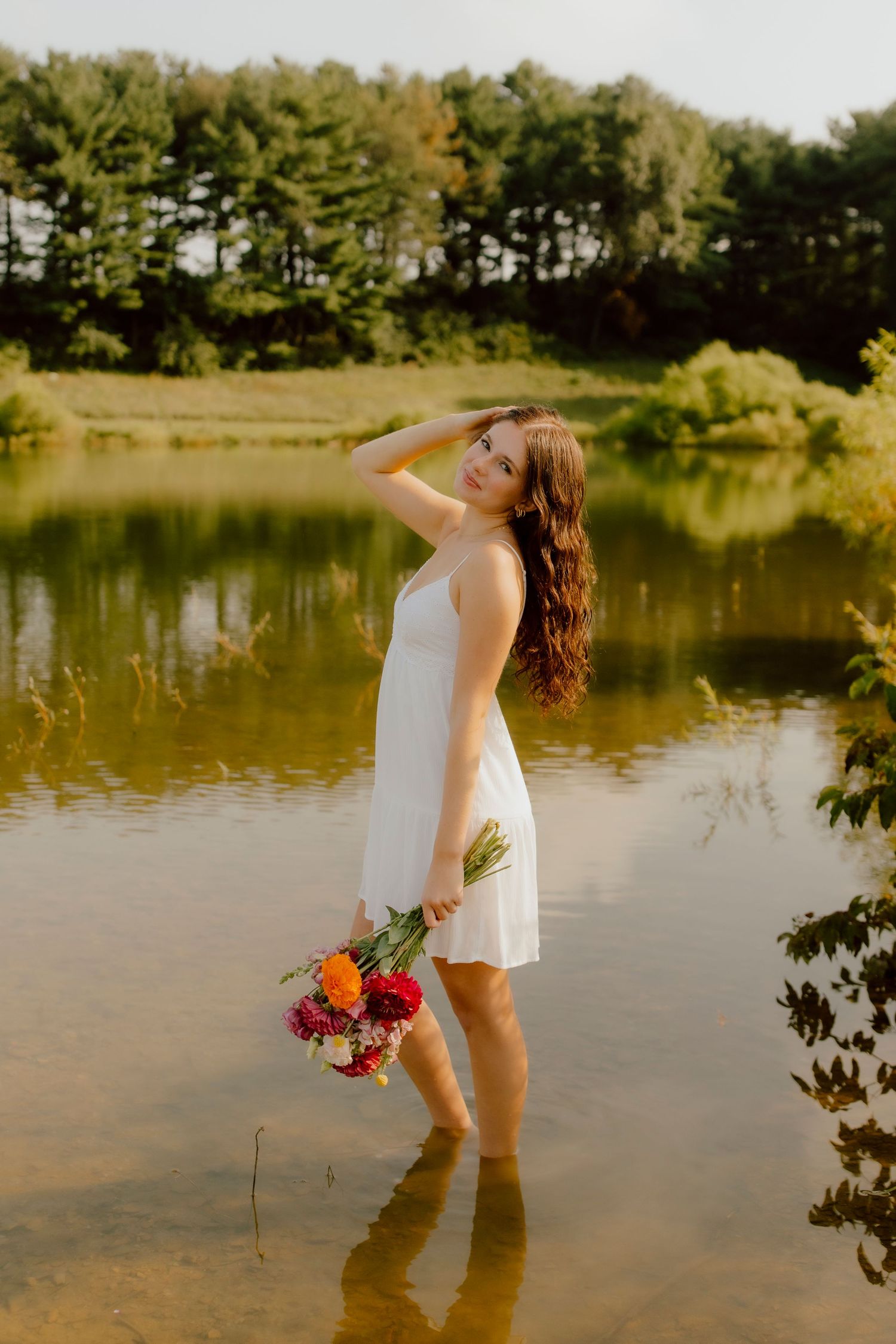 Young woman in white dress standing in shallow water holding colorful bouquet with forest background.