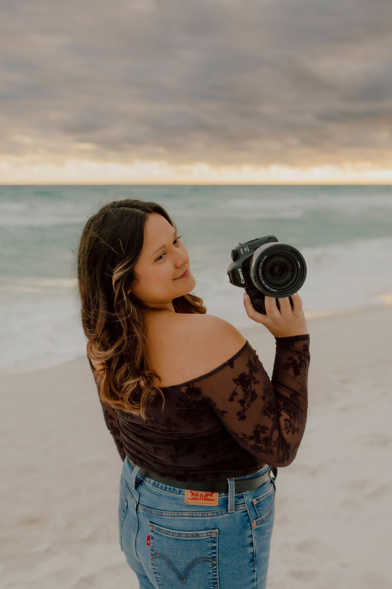 Young female photographer smiling while holding a DSLR camera on a beach at sunset.
