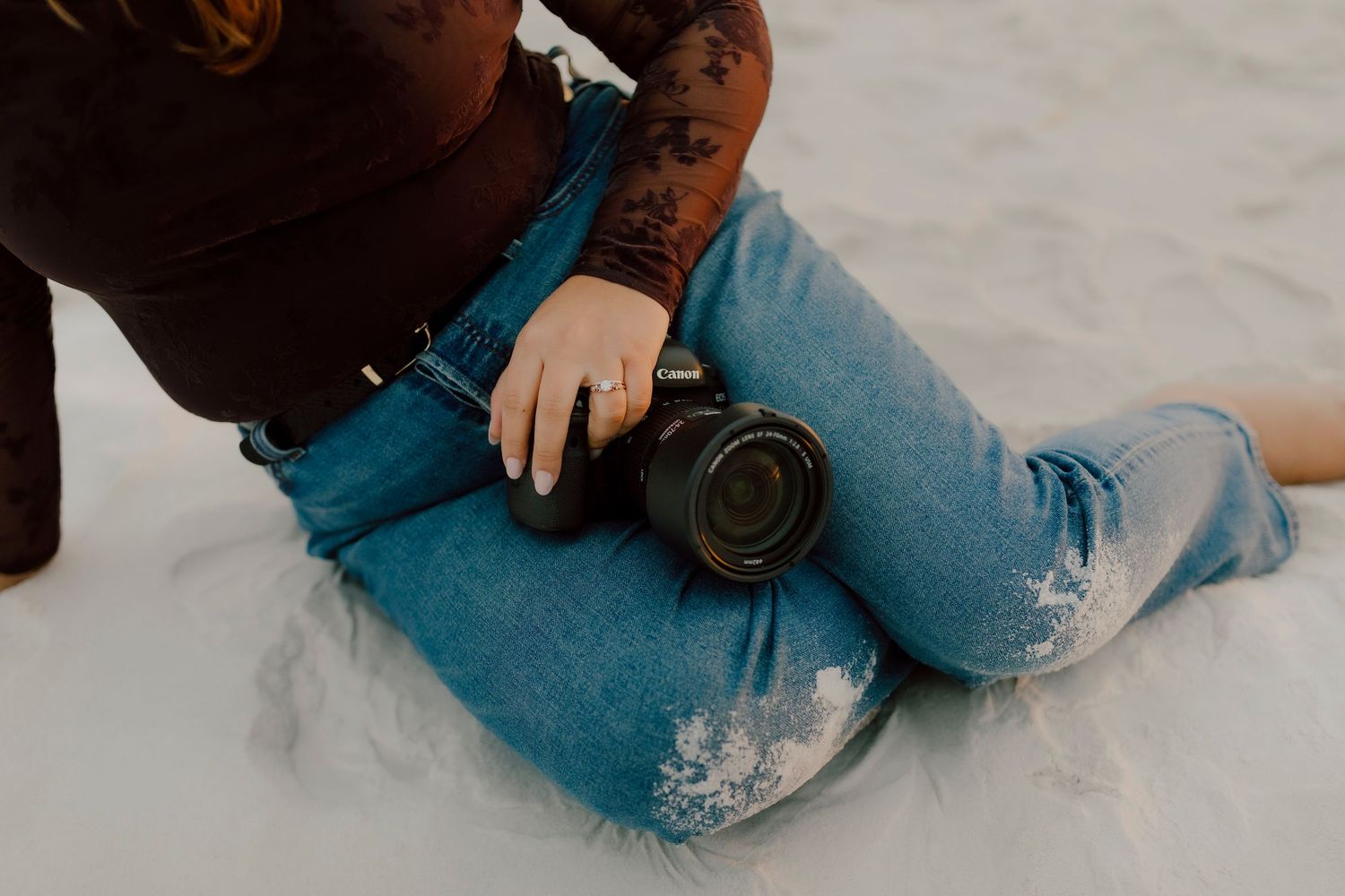 Person with tattoos sitting on sand, holding a black camera, wearing blue jeans and a brown top.