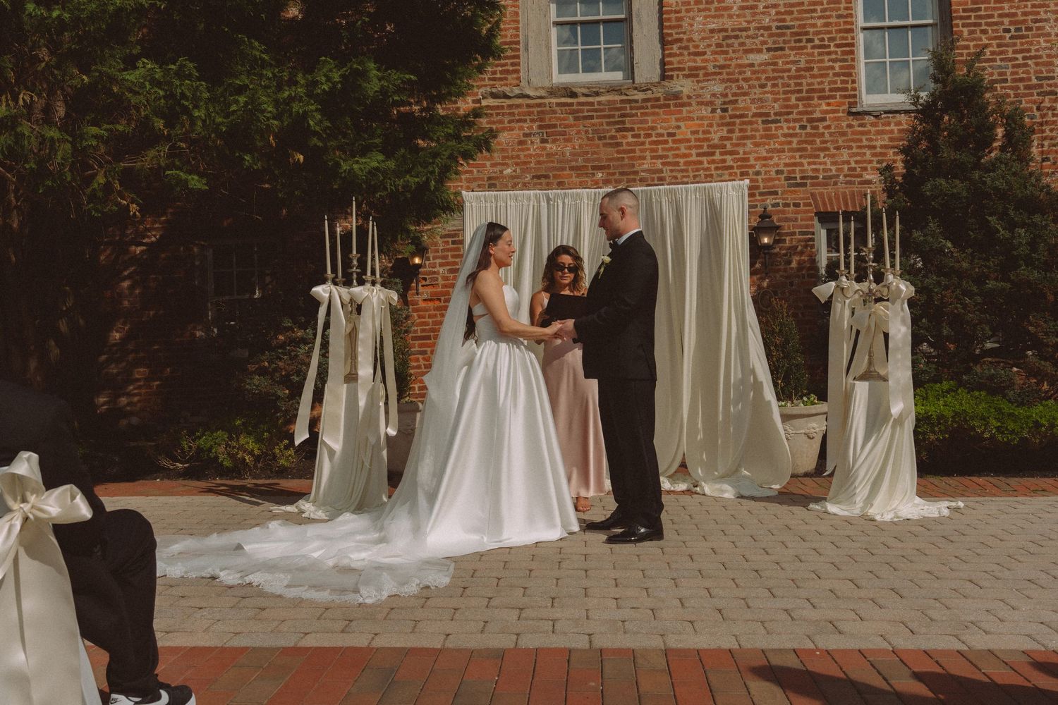 Outdoor wedding ceremony at brick venue with draped arch, bride in white gown, groom, and officiant on brick patio.