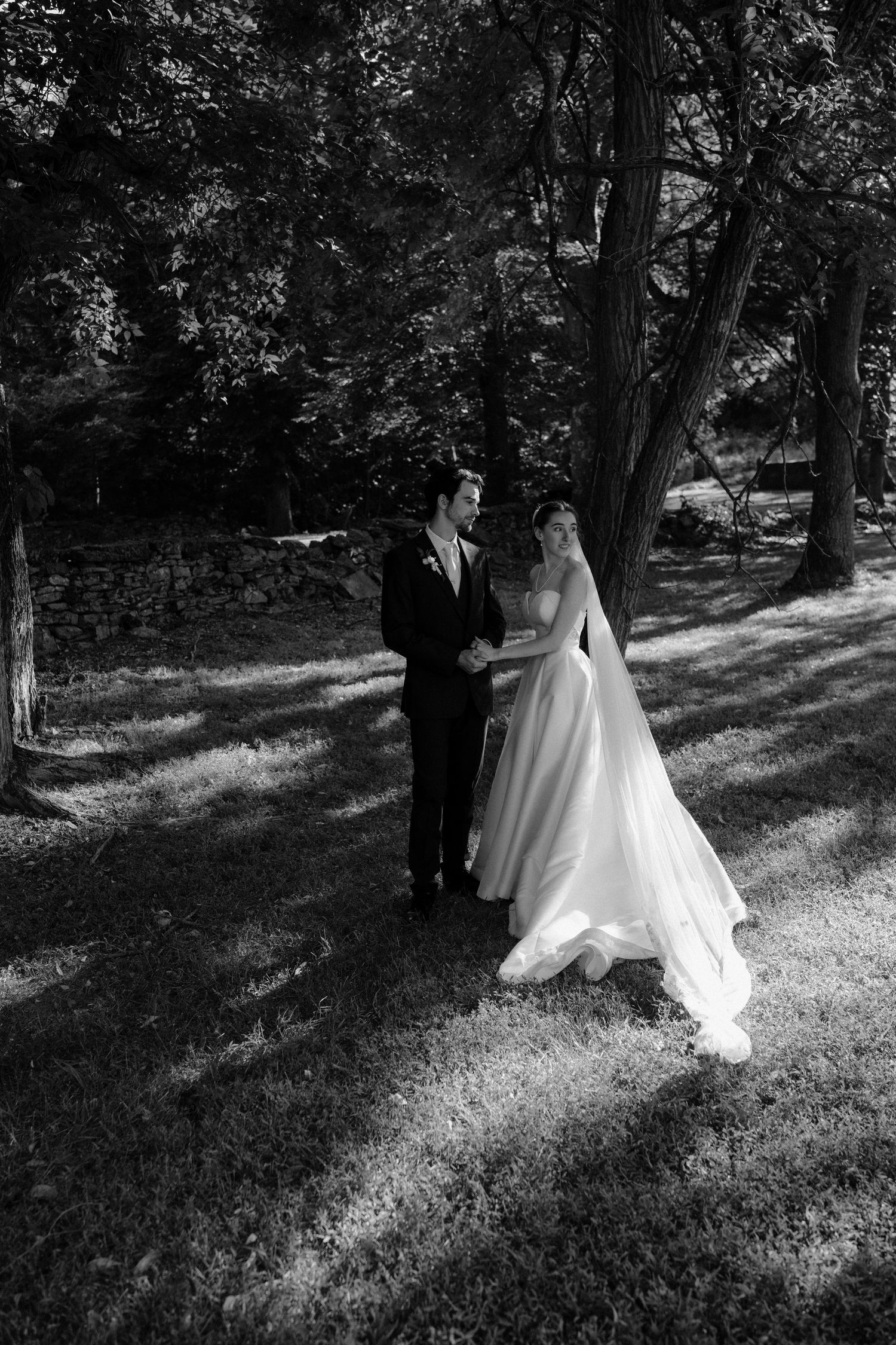 Black and white photo of a bride and groom holding hands in a sunlit forest, wearing wedding attire.