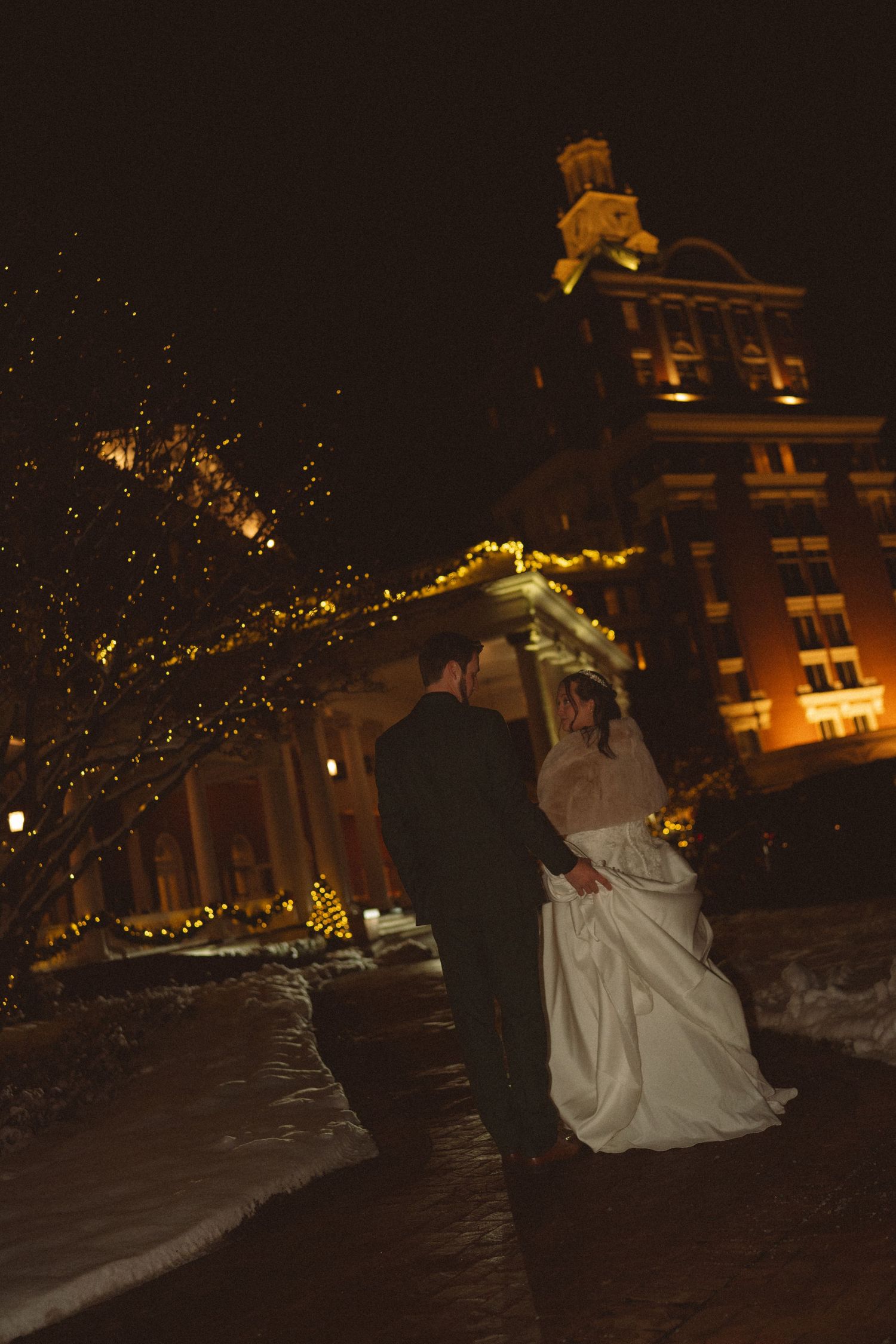 Bride and groom share a romantic moment outdoors at night, surrounded by twinkling lights and a historic building backdrop.