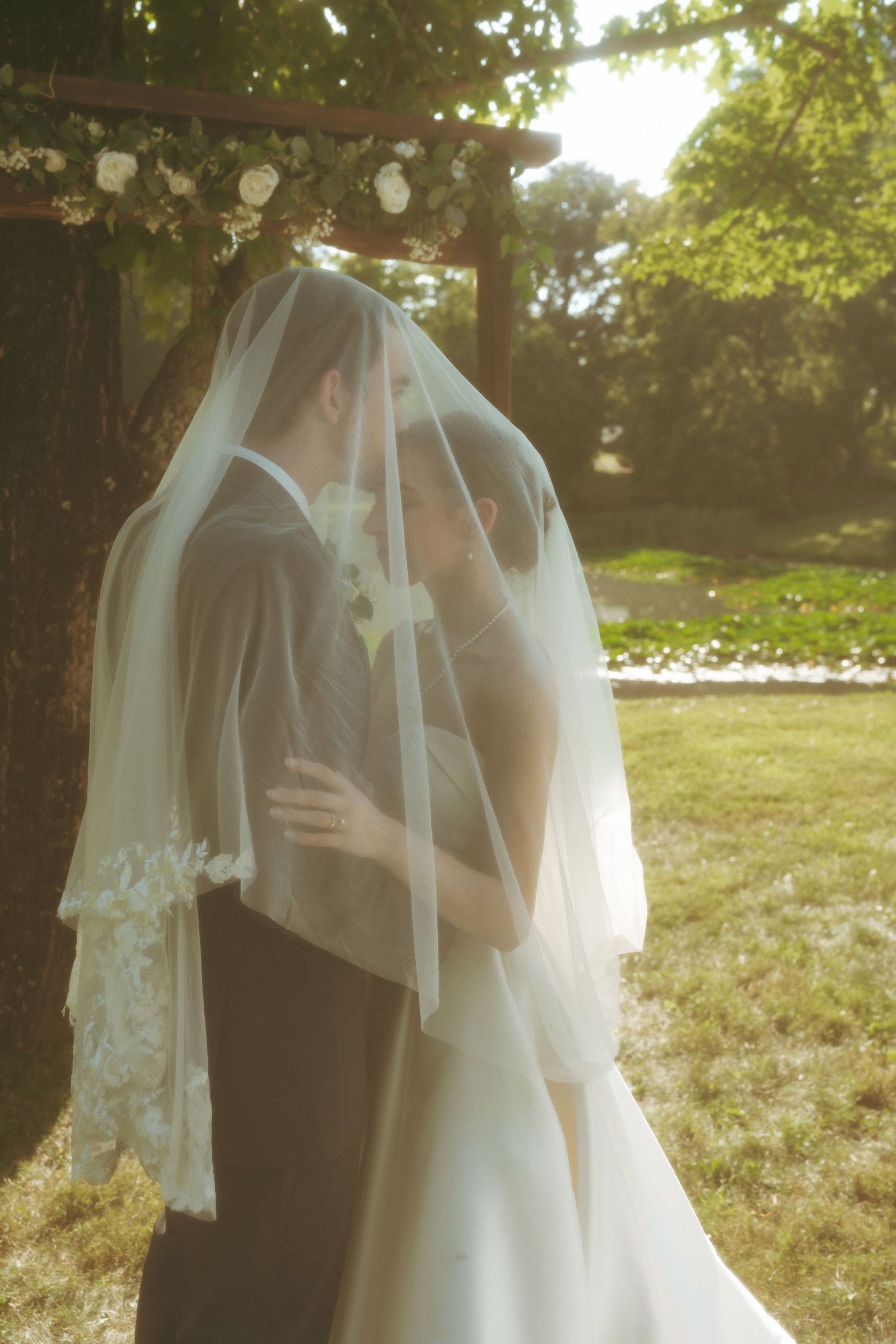 Bride and groom sharing a kiss under a white veil at an outdoor wedding ceremony surrounded by lush greenery.
