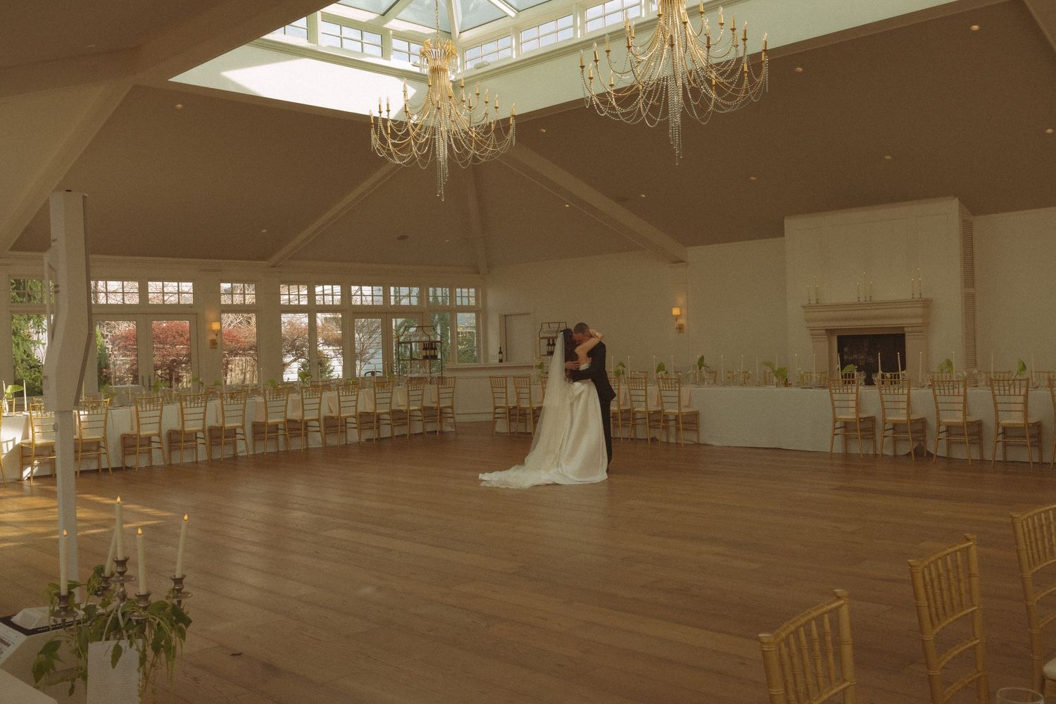 Bride and groom share first dance in elegant wedding venue with chandeliers, gold chairs, and hardwood floors.