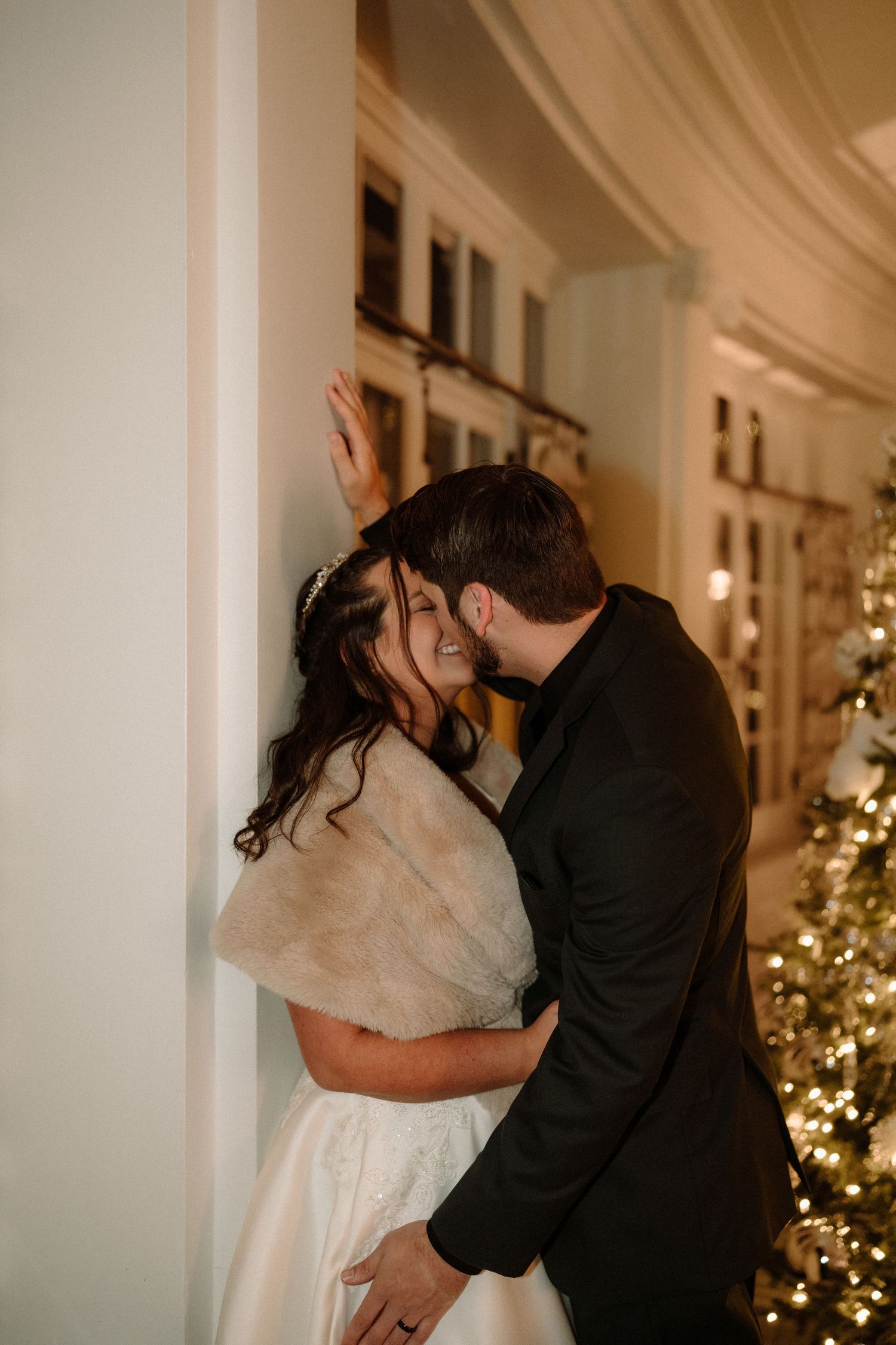 Bride in white gown and fur wrap smiles as groom kisses her cheek by a Christmas tree.