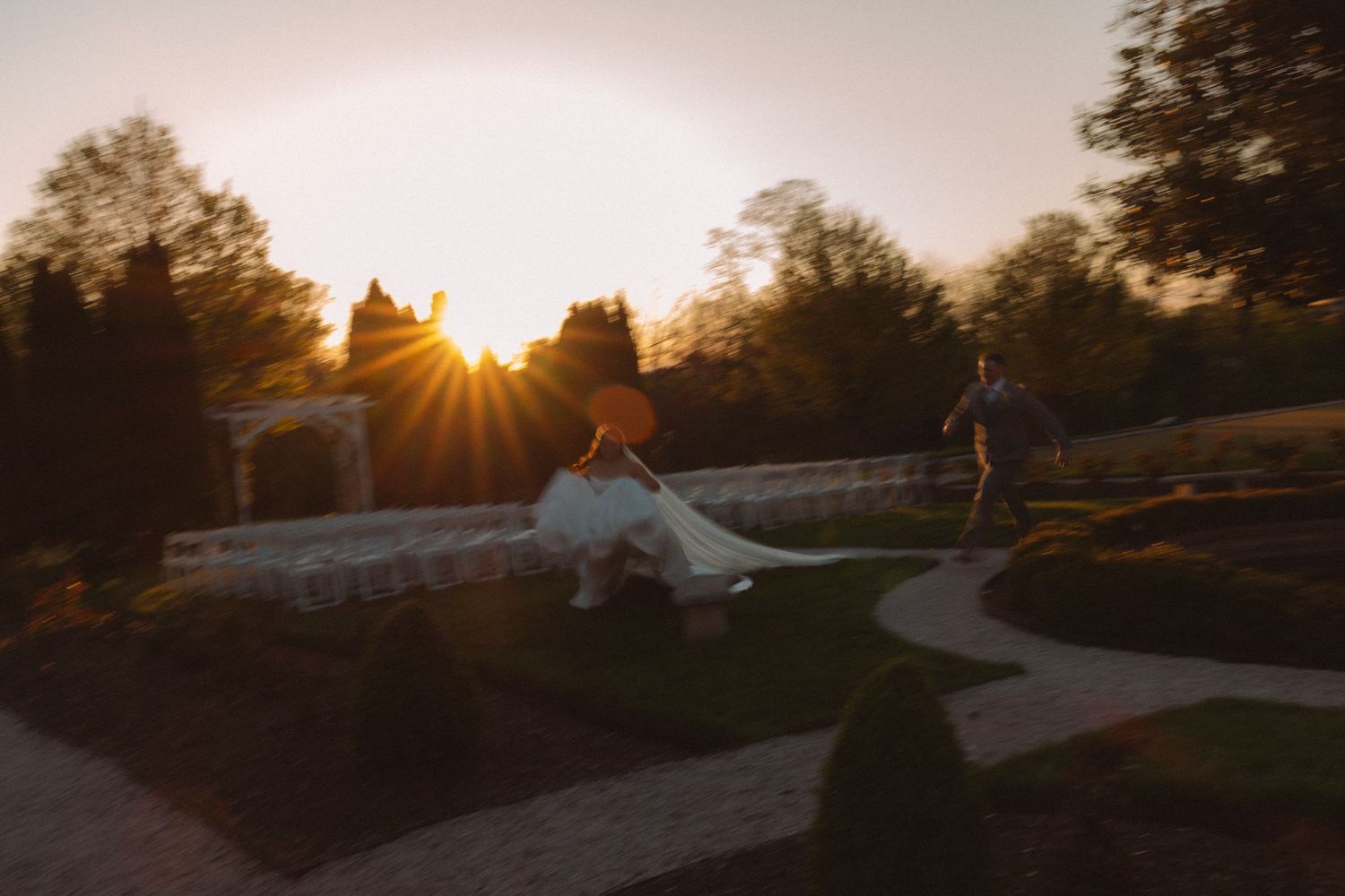 Bride and groom walk through a formal garden at golden sunset, wedding dress trailing elegantly on lush green path.