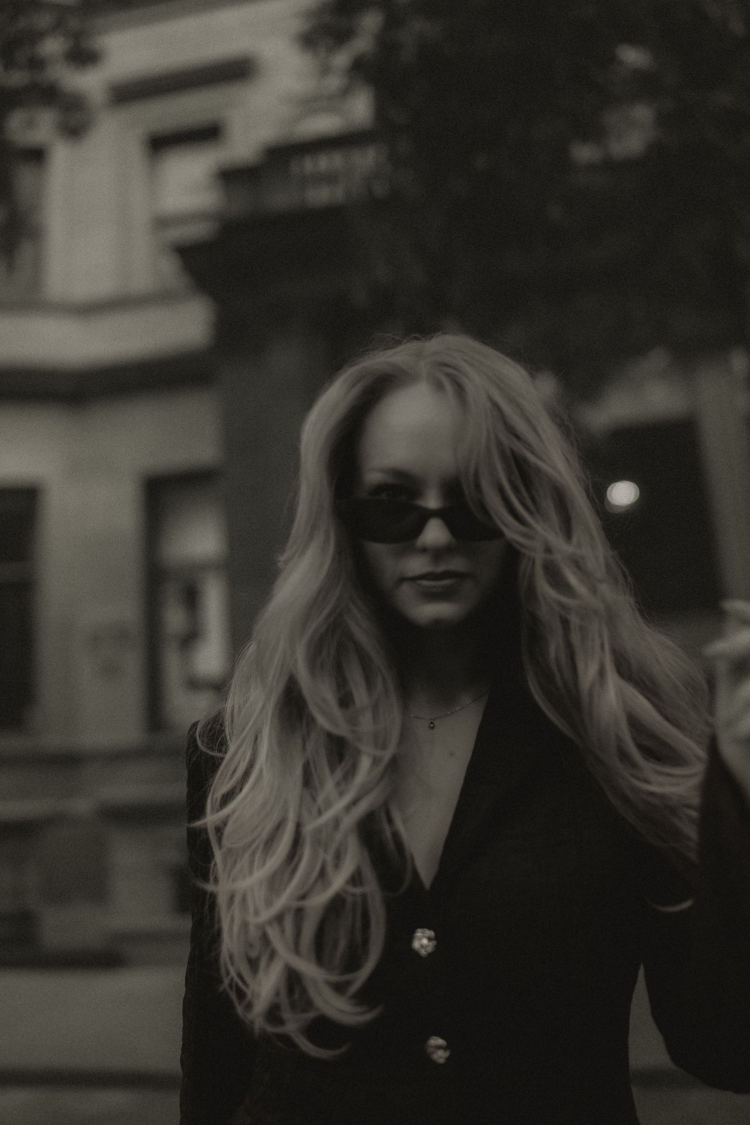 Blonde woman in black blazer and sunglasses poses outdoors in moody black and white street photography.