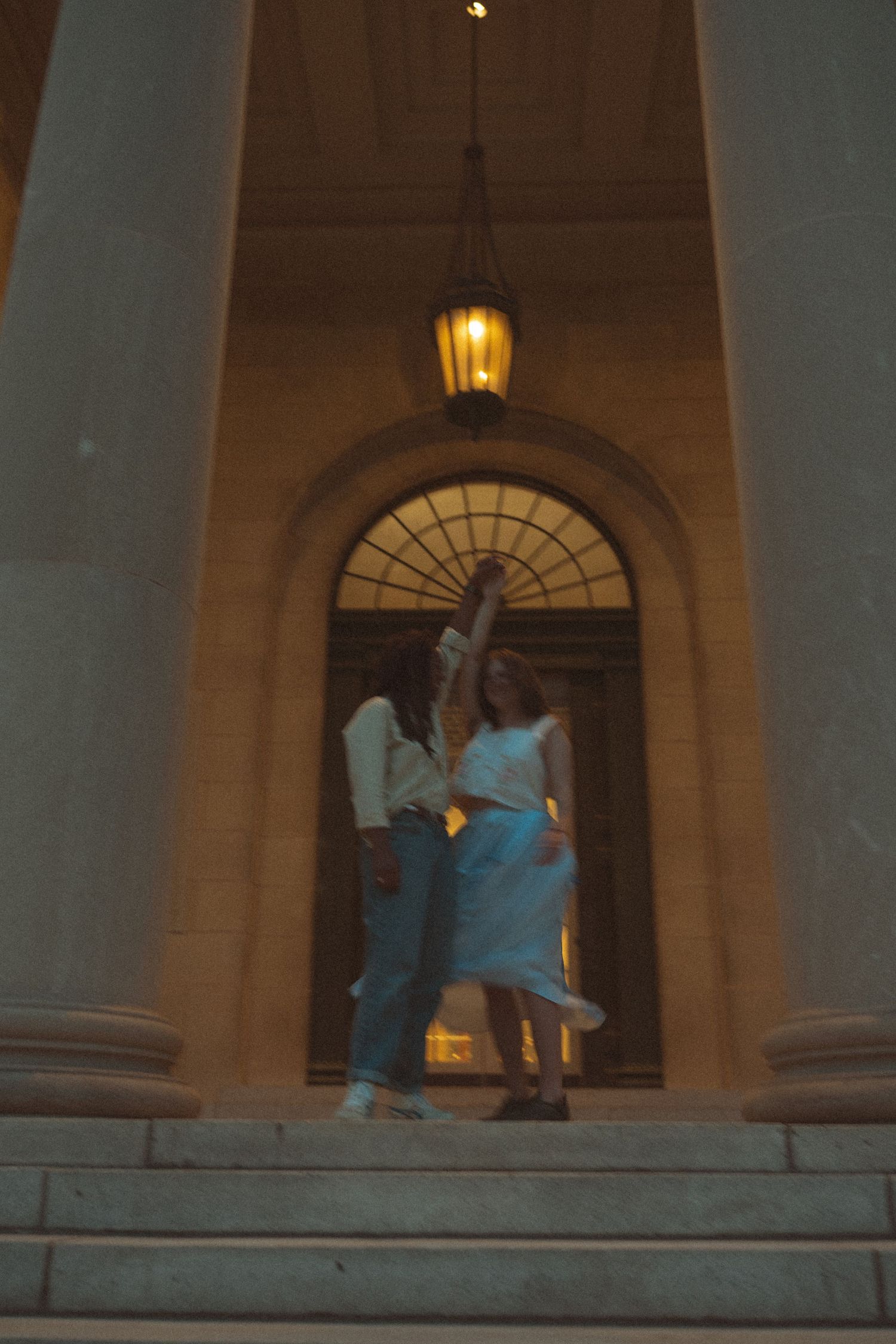 Couple dancing on grand building steps at night, illuminated by warm lantern light under an arched doorway.