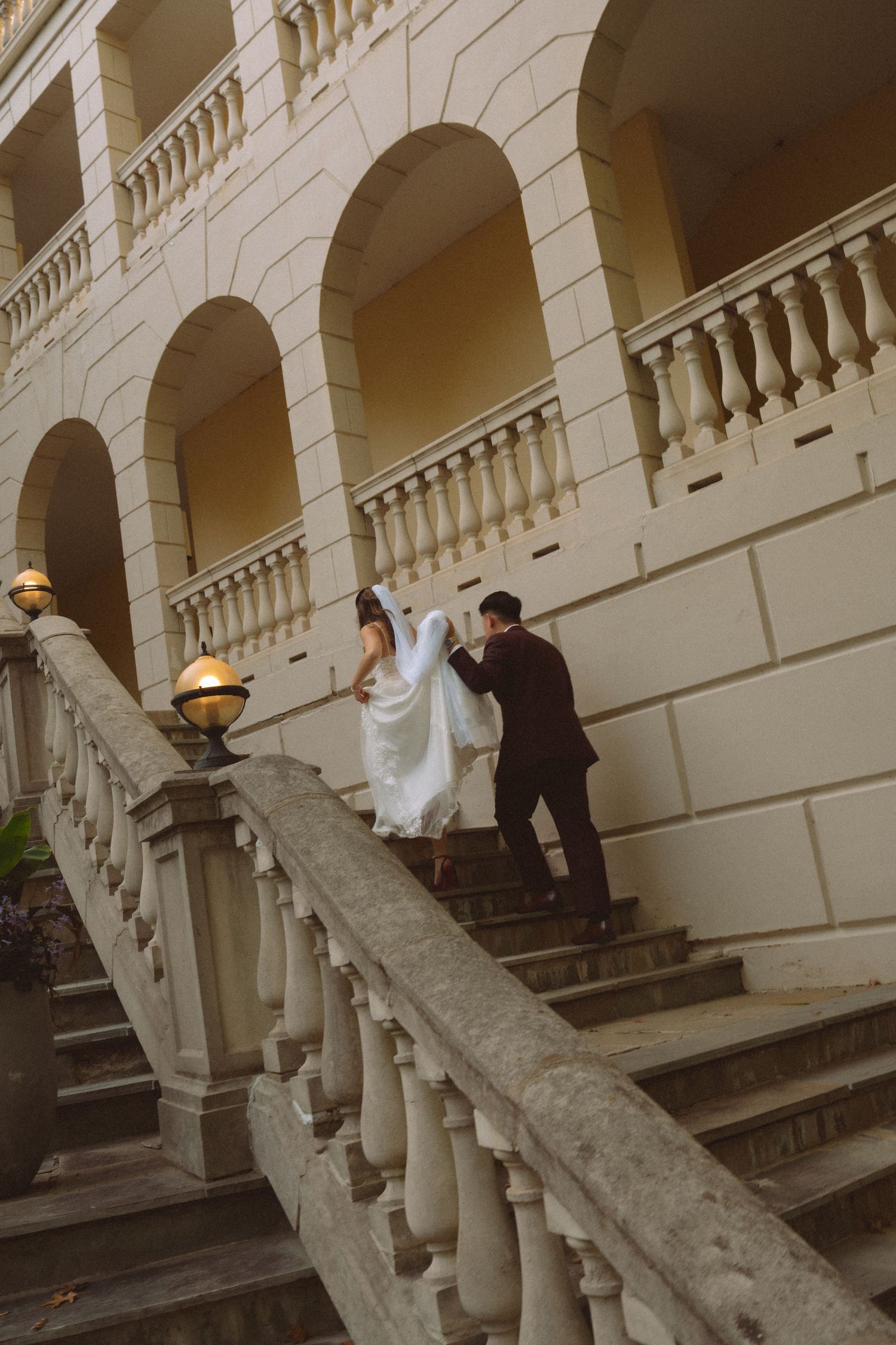 Bride and groom ascending elegant stone staircase with arched colonnade at wedding venue.