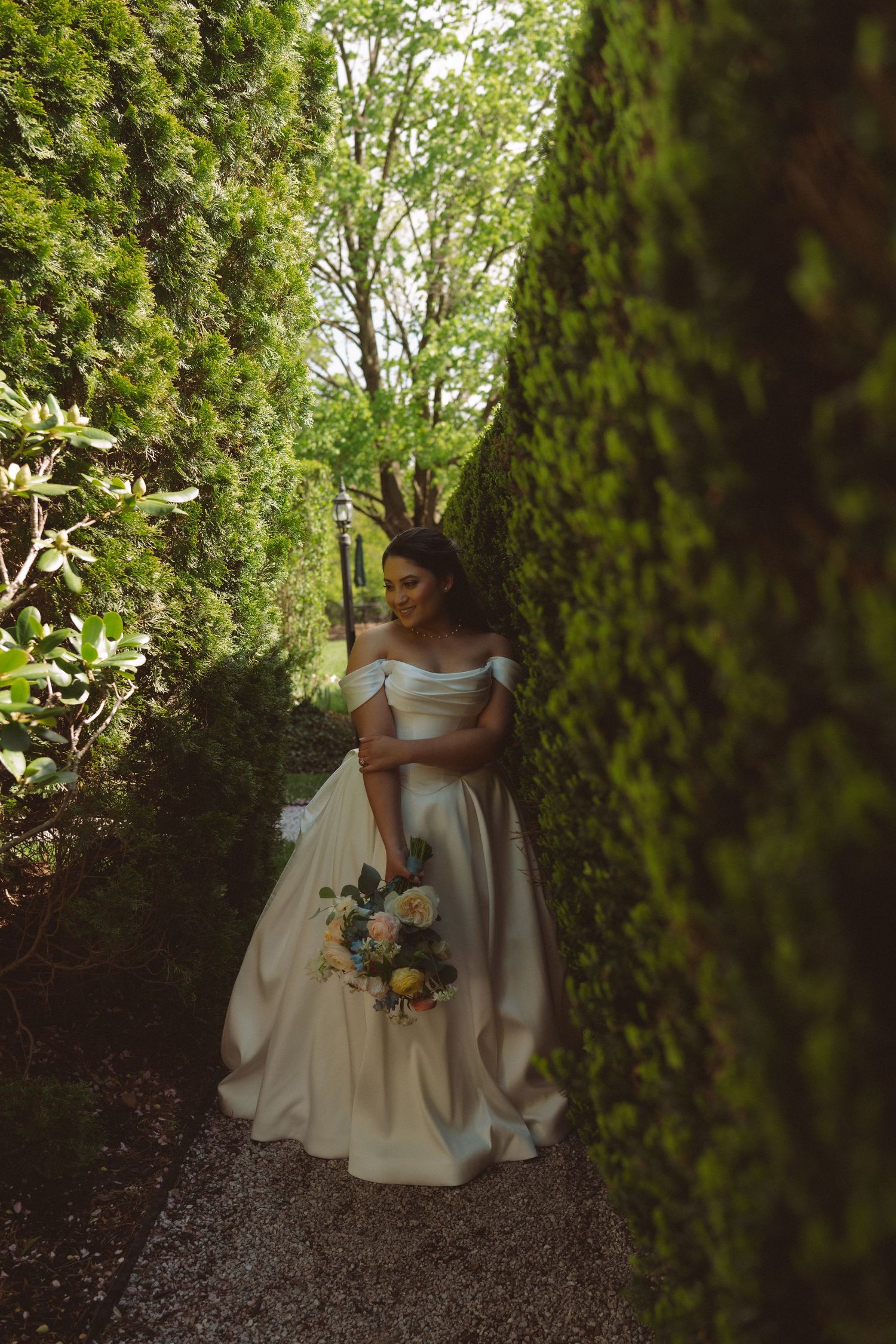 Bride in off-shoulder white gown holds bouquet in lush garden hedge pathway.