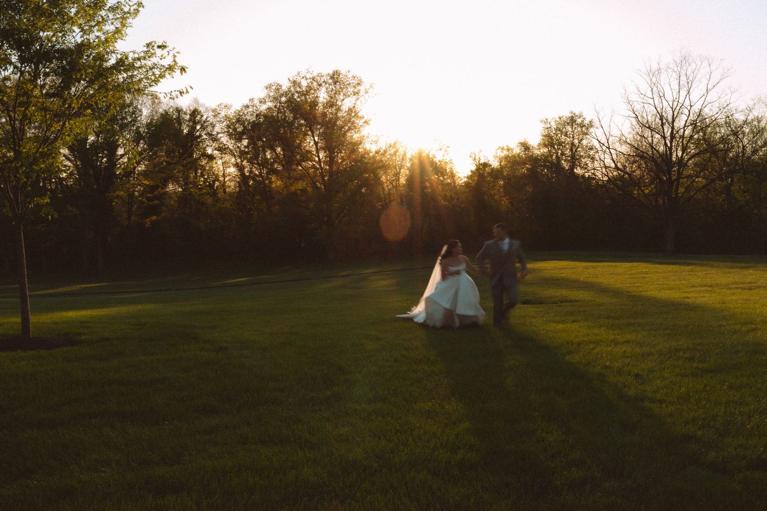 Bride and groom walk hand in hand across lush green lawn at golden hour sunset with warm sunlight glowing through trees.