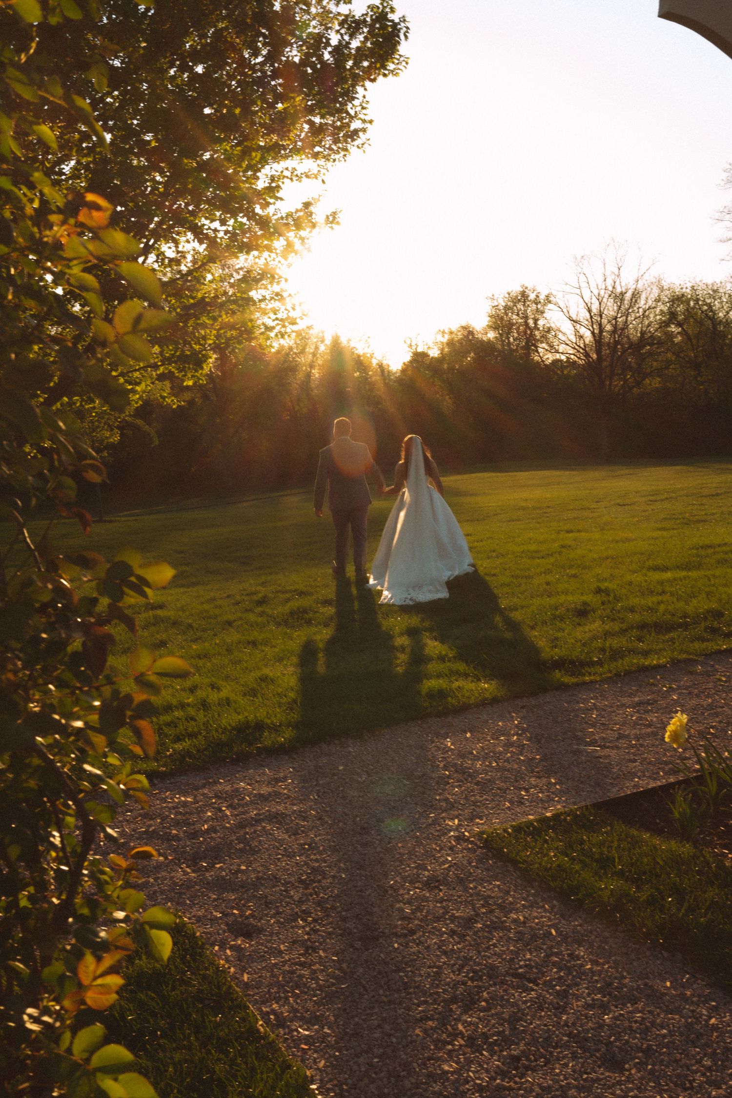 A bride and groom walk hand in hand along a garden path at golden sunset, casting long shadows on lush green grass.