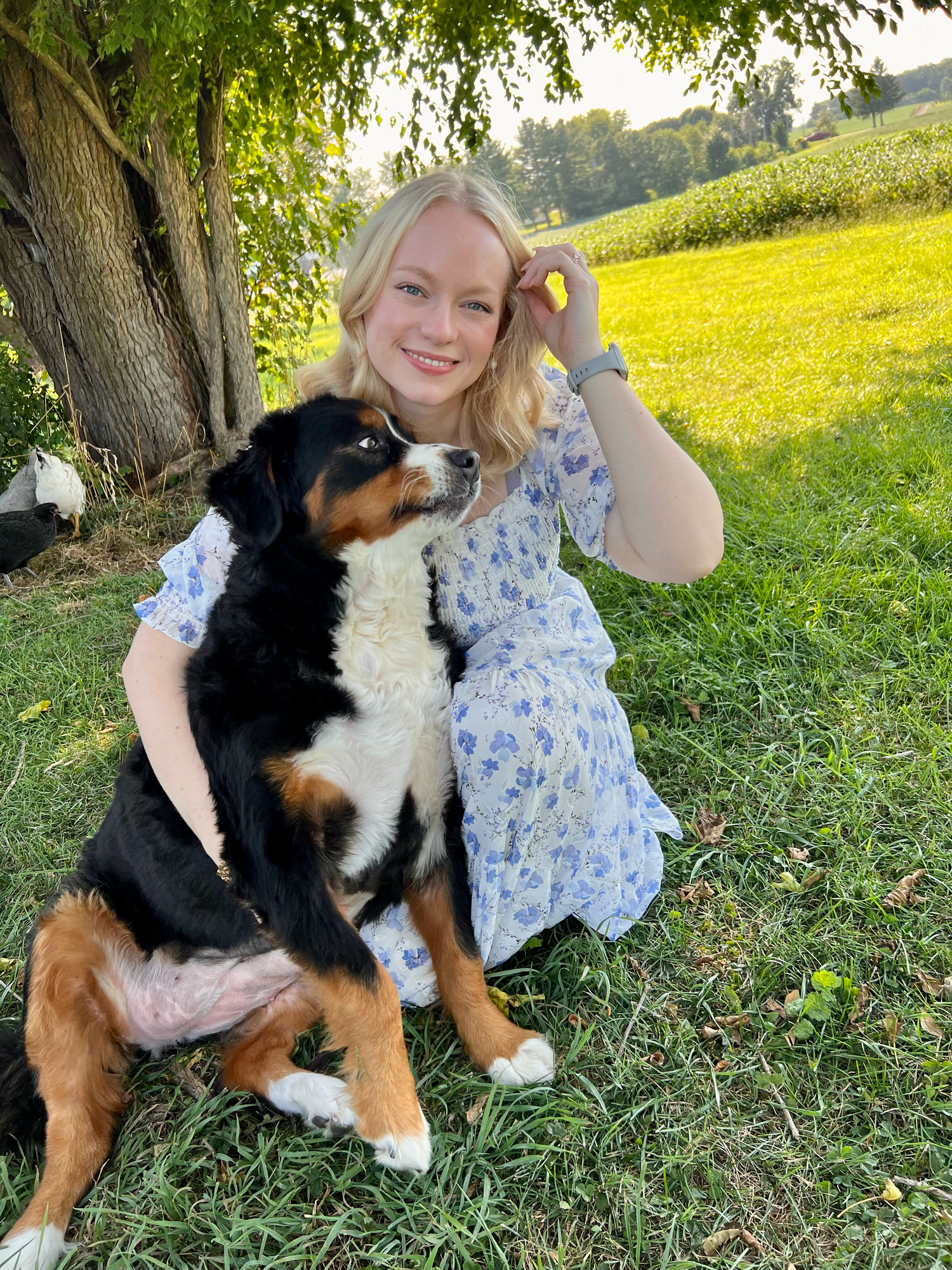 Young blonde woman in floral dress smiling with Bernese Mountain Dog puppy on green grass near tree.