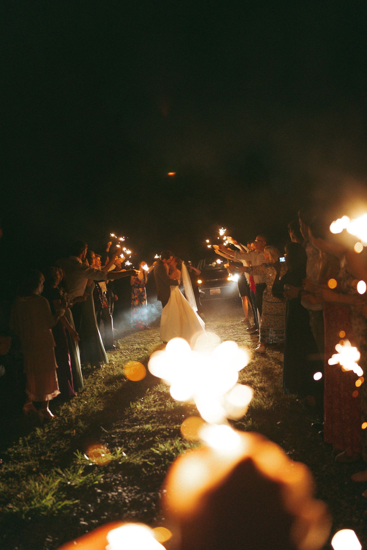 Bride walks through sparkler tunnel at night wedding send-off with guests lining the path holding glowing sparklers.