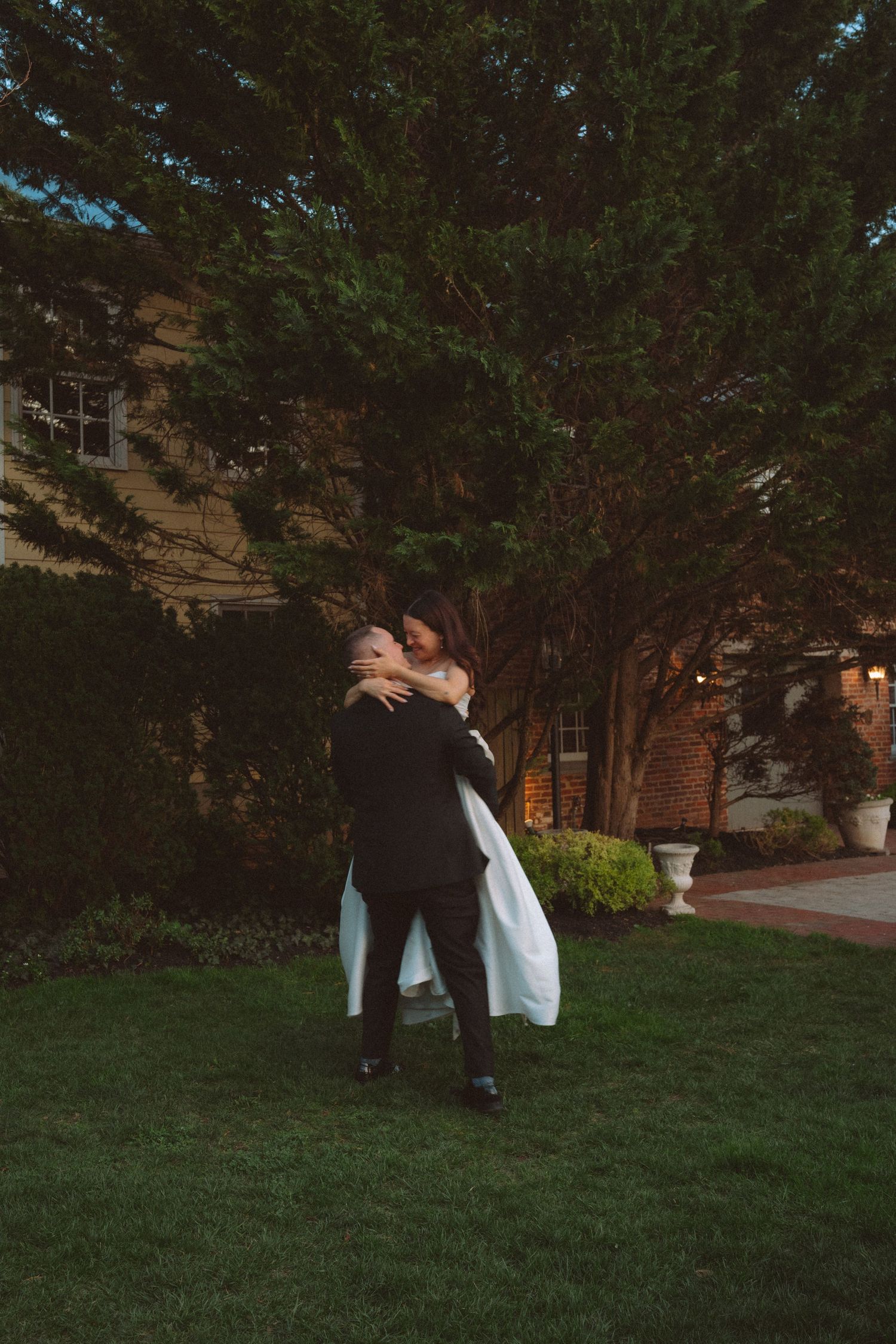 Groom lifting bride in white gown outdoors at dusk, embracing near lush green trees at elegant wedding venue.