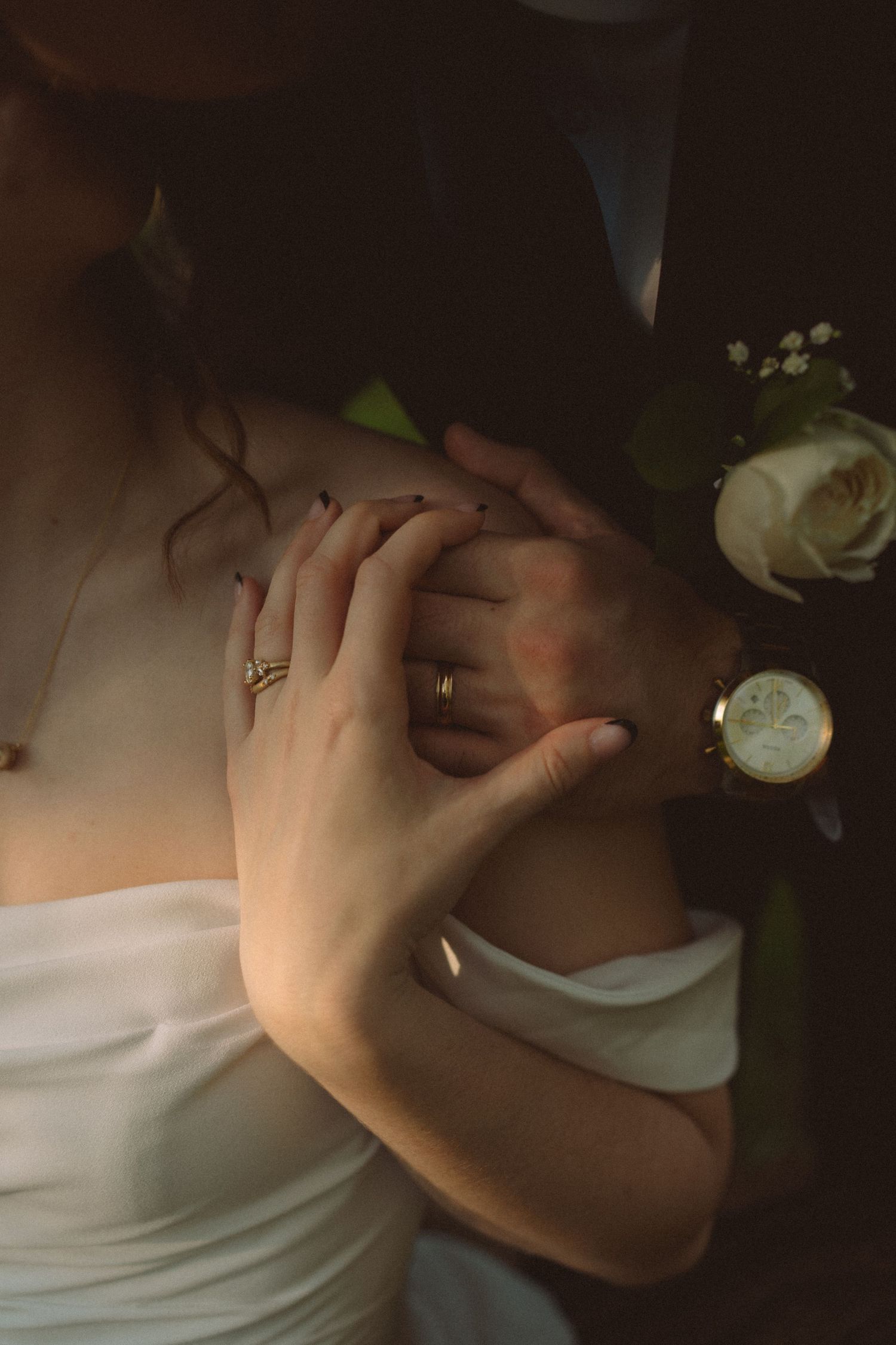 Close-up of bride and groom hands with wedding rings, watch, and white rose boutonniere in warm light.