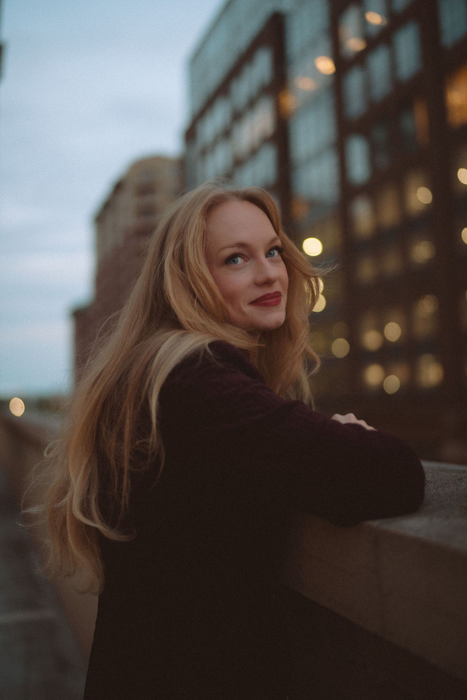 Young blonde woman with red lips smiles over shoulder on urban rooftop at dusk, city lights bokeh background.