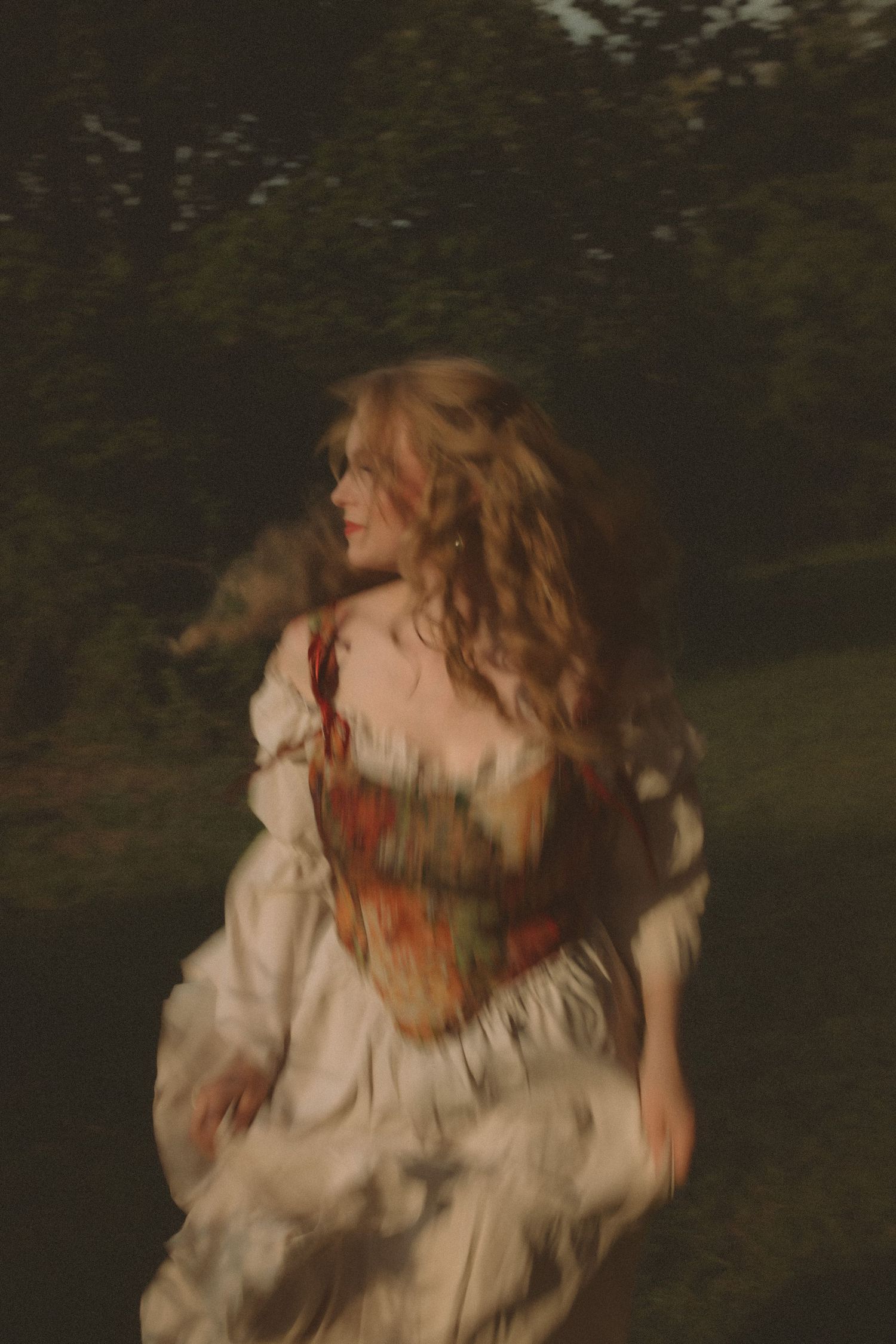 A blurred woman in a floral corset and white dress twirls in a dark garden, her curly hair flowing.
