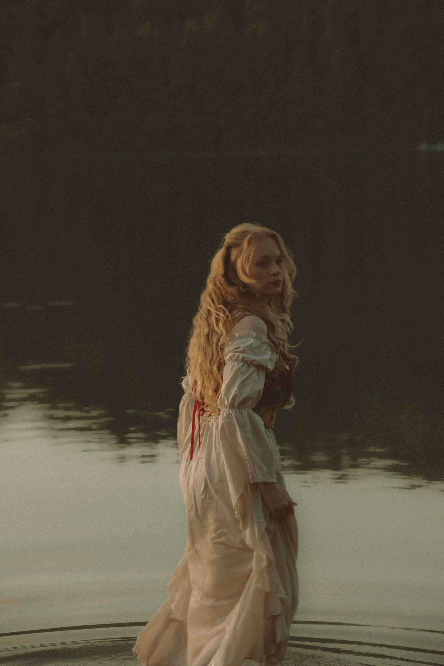 A woman in a vintage white dress with red accents stands in a misty lake, her long blonde hair flowing behind her.