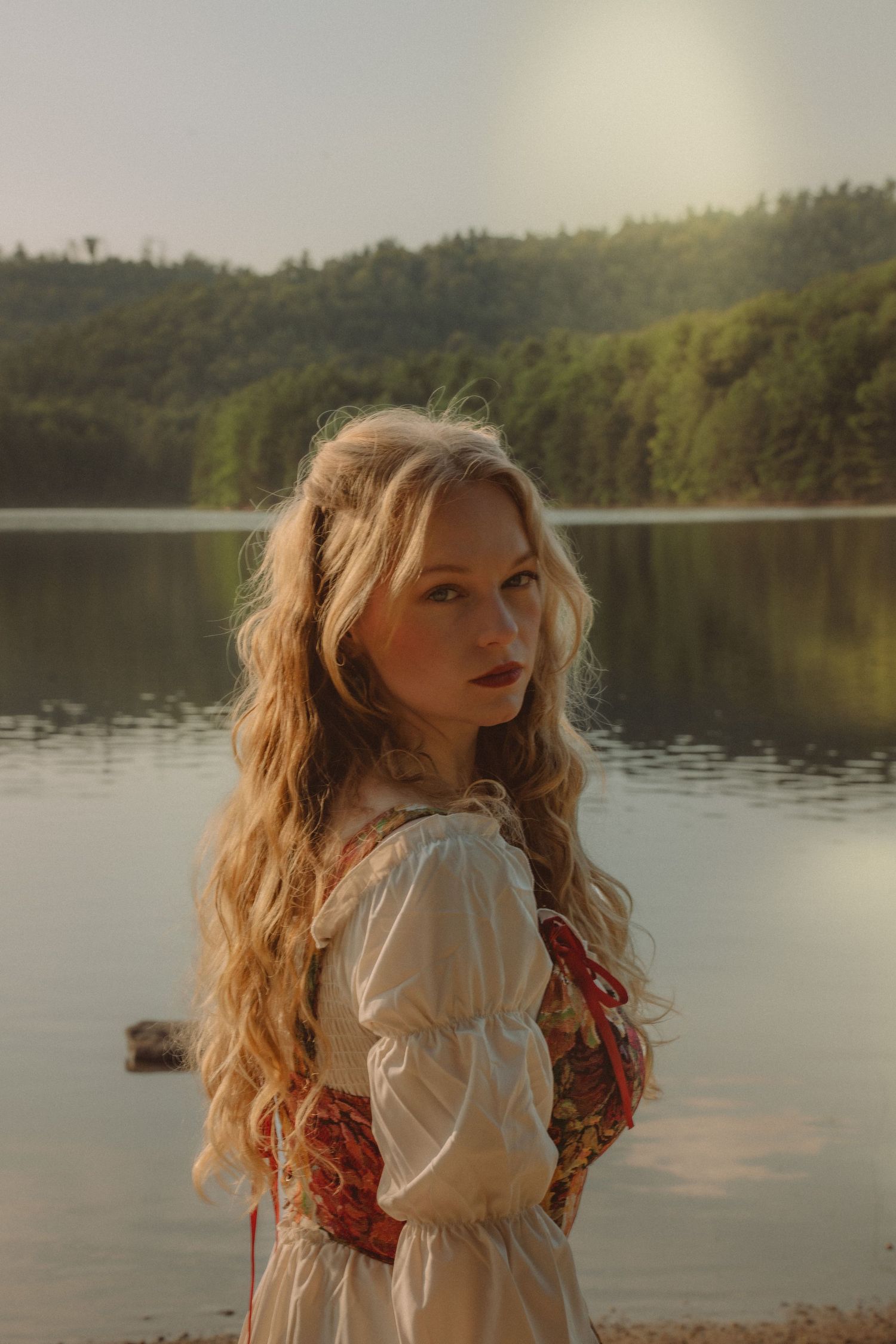 Young woman with long blonde wavy hair in vintage floral dress poses by a serene lake at golden hour.