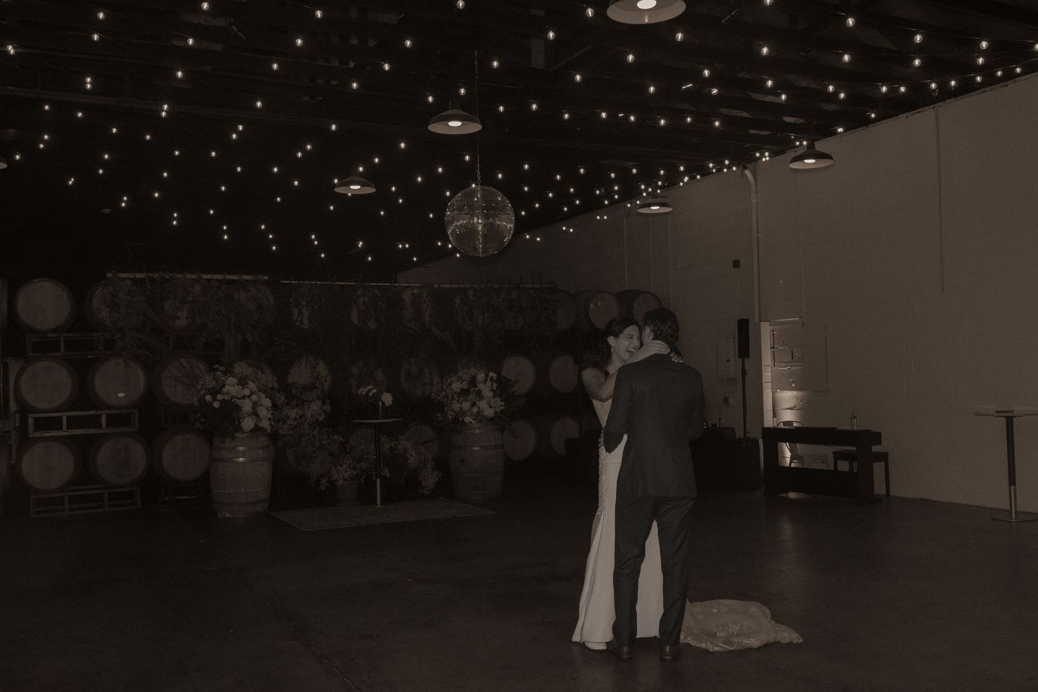 Couple shares first dance under twinkling fairy lights and disco ball in rustic winery venue with barrel backdrop.